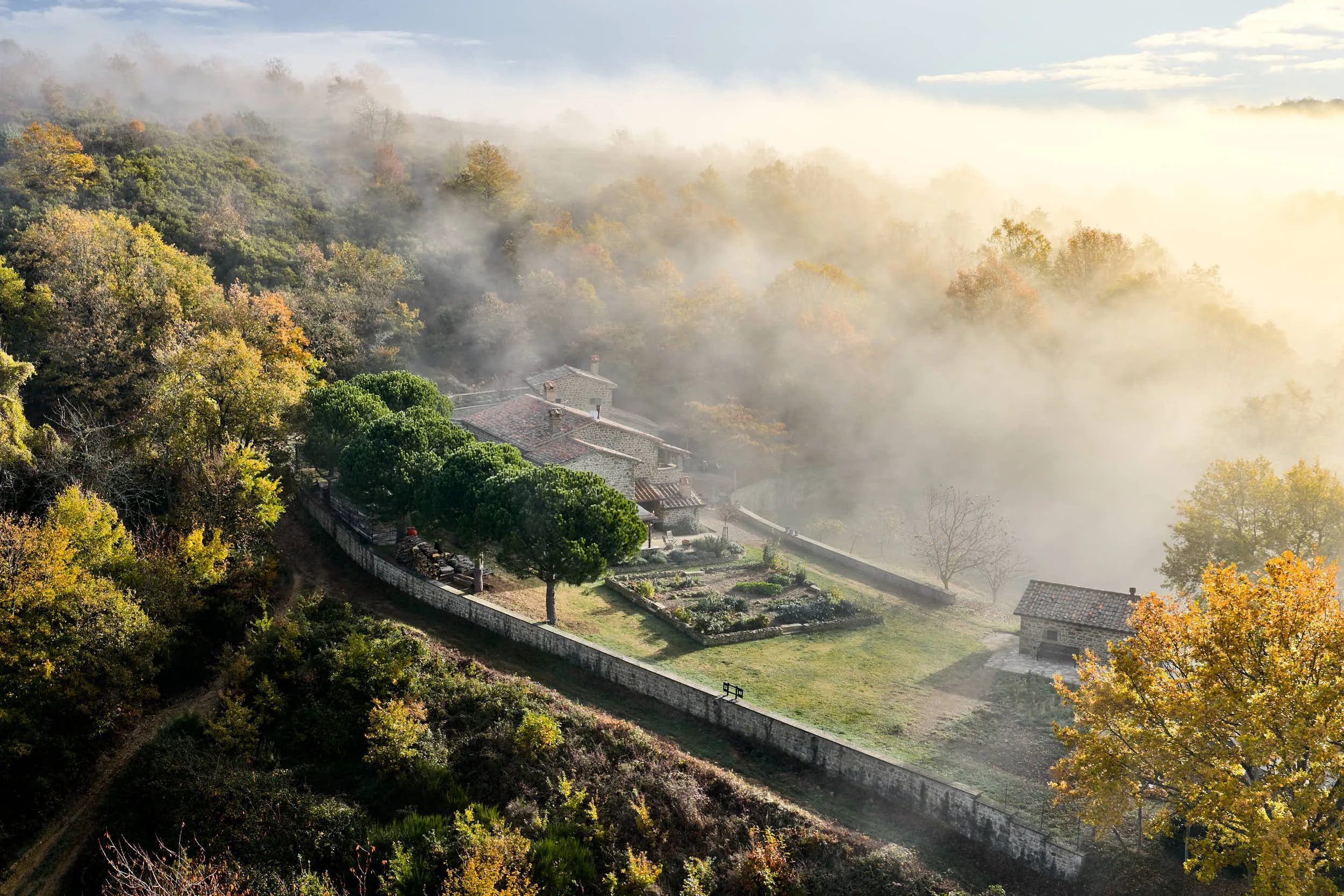 Ein Haus inmitten eines bewaldeten Hügels, umgeben von Bäumen und einem stone wall, im Nebel morgens bei Sonnenaufgang.