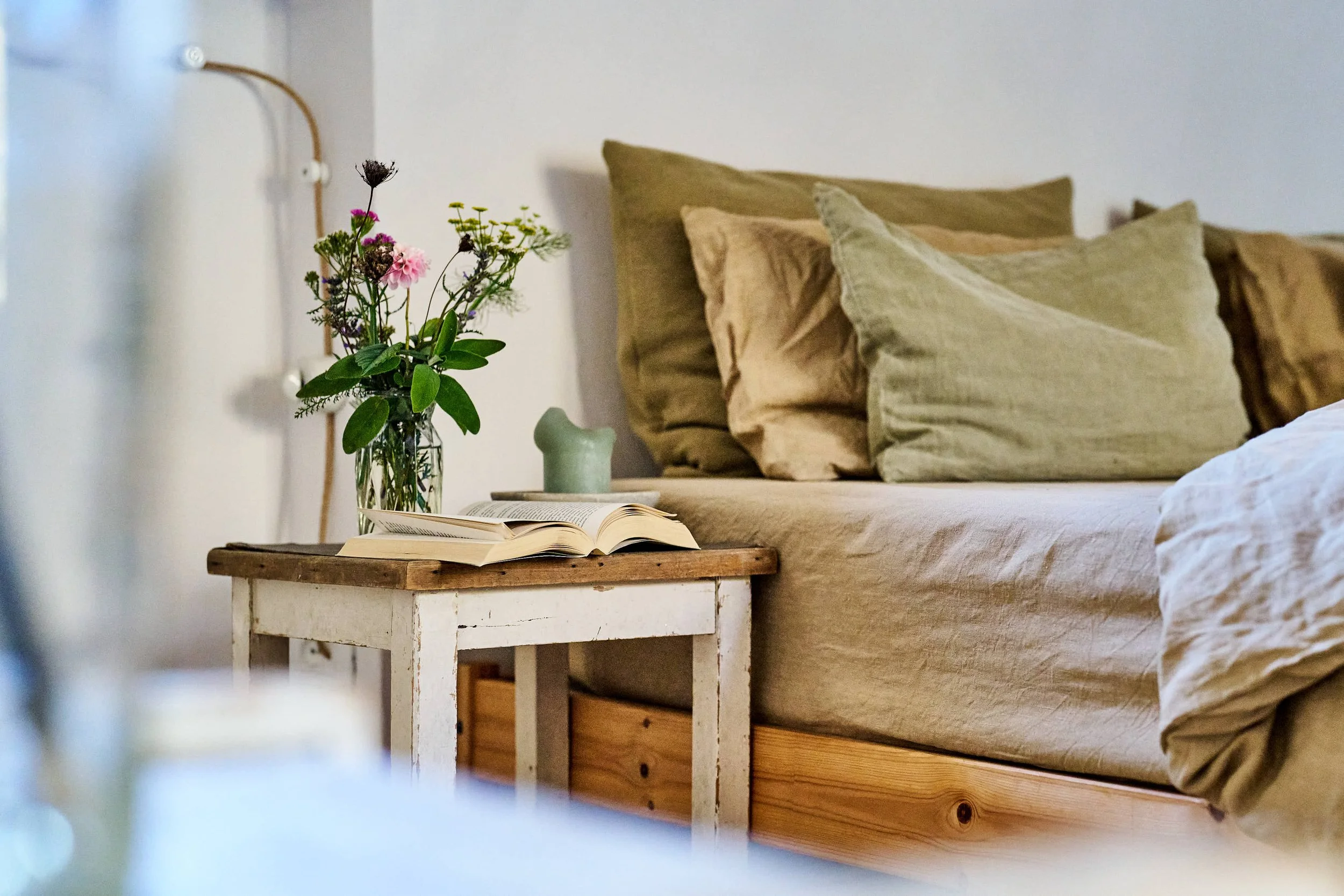 Bett mit grünen Kissen, Nachttisch mit offenem Buch und Blumenstrauß in Glasvase.