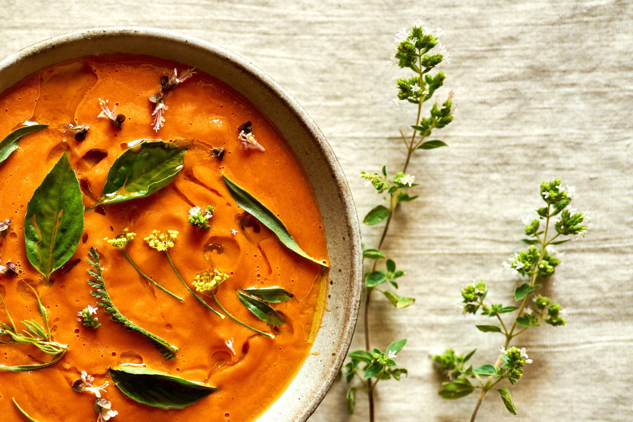 Eine Schüssel mit einer orangefarbenen Suppe, garniert mit frischen grünen Blättern und kleinen Blumen, neben einem Zweig mit kleinen weißen Blüten auf einem hellen Stoffhintergrund.