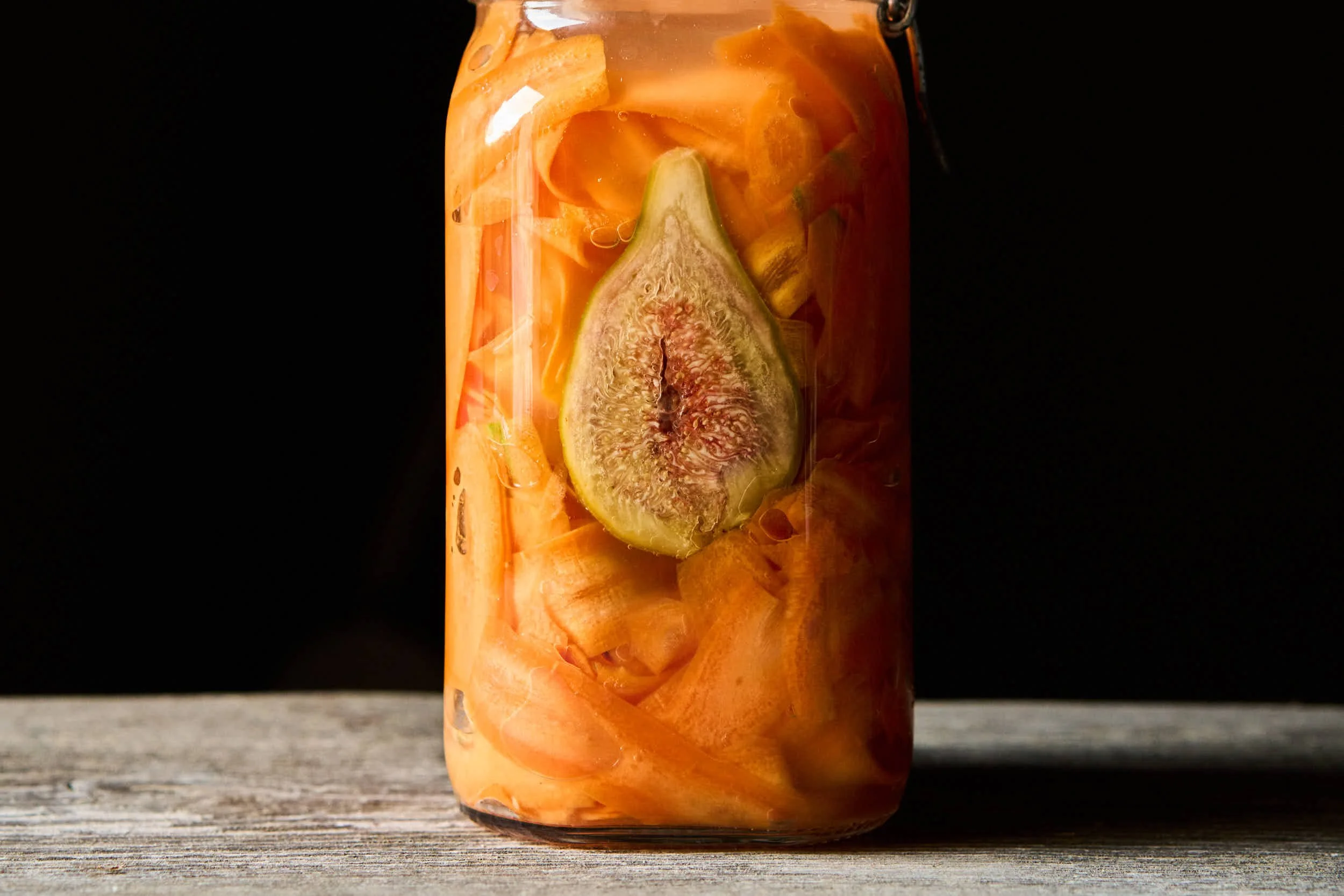 Ein Glas mit eingelegten Feigen, Karotten und anderem Gemüse in einer klaren Flüssigkeit, auf einer hölzernen Oberfläche vor einem schwarzen Hintergrund.