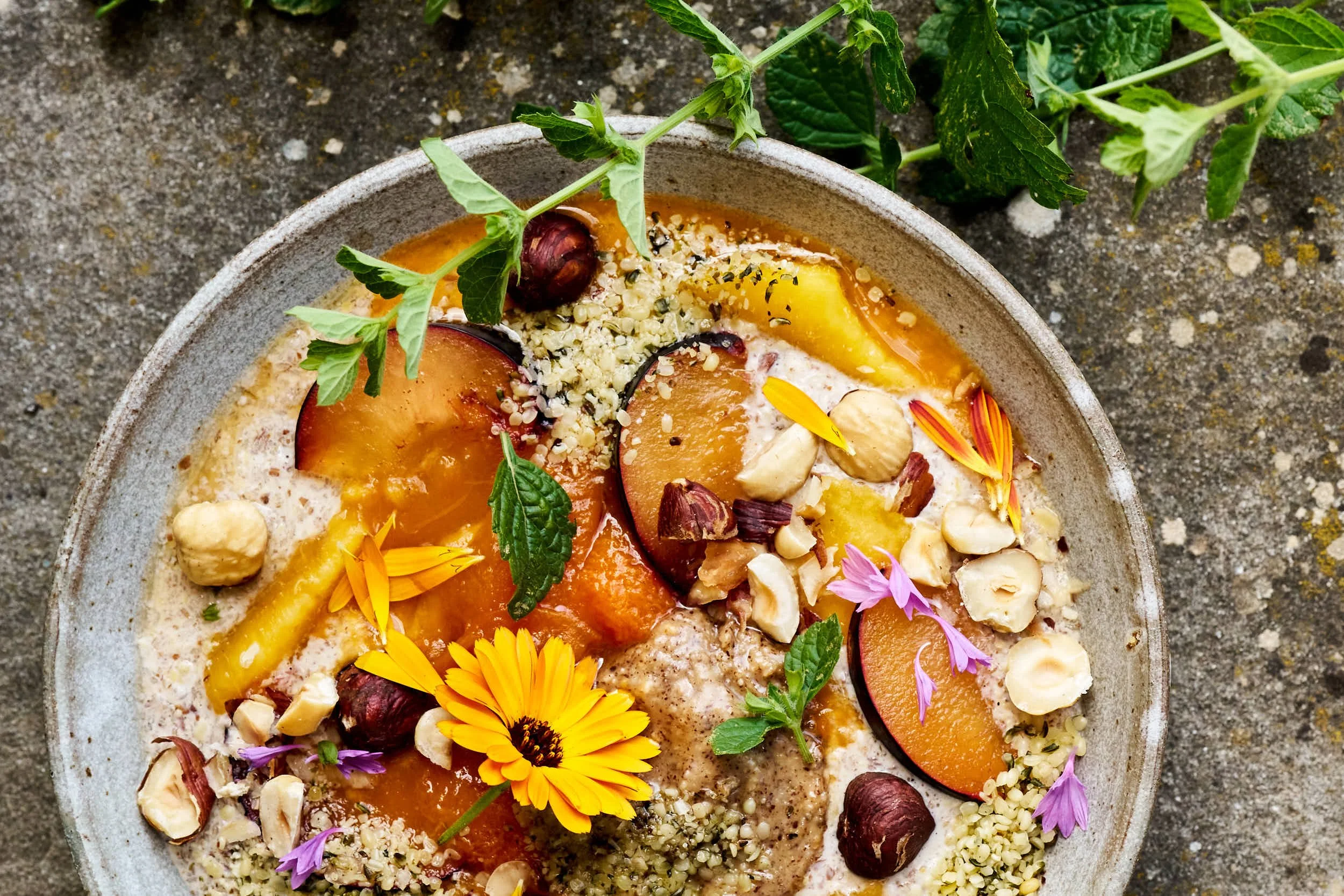 Bunter smoothie bowl mit Pfirsichen, Nüssen, Blüten und Minzblättern auf einem grauen Steinuntergrund.