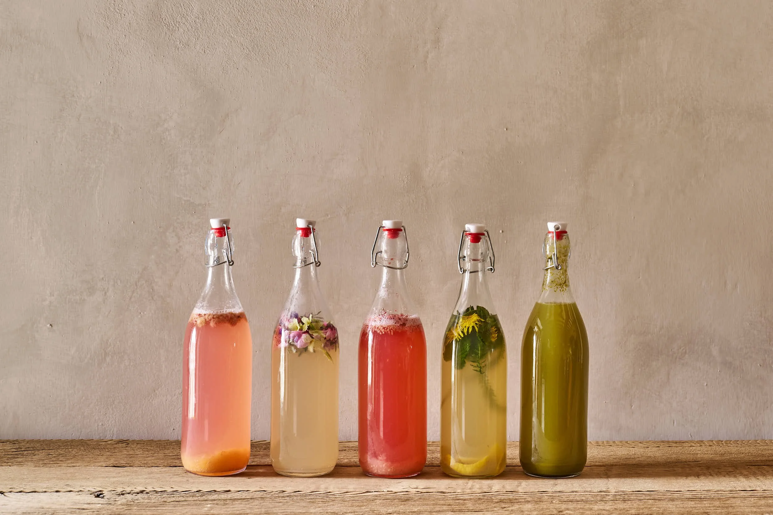Fünf Glasflaschen mit bunten, gefüllten Getränken und Kräutern auf einem Holztisch gegen eine helle Wand