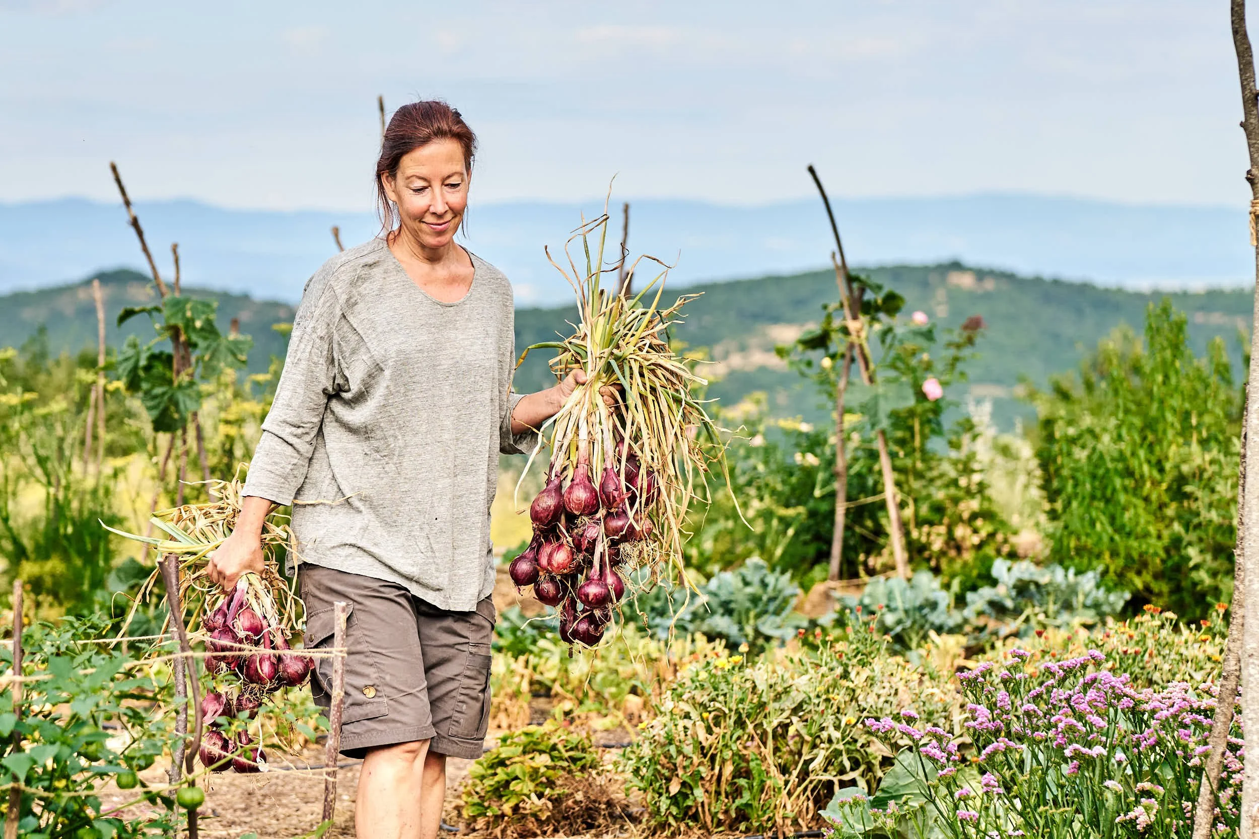 Eine Frau erntet knoblauch in einem Garten mit Gemüse und Blumen, im Hintergrund sind Berge zu sehen.