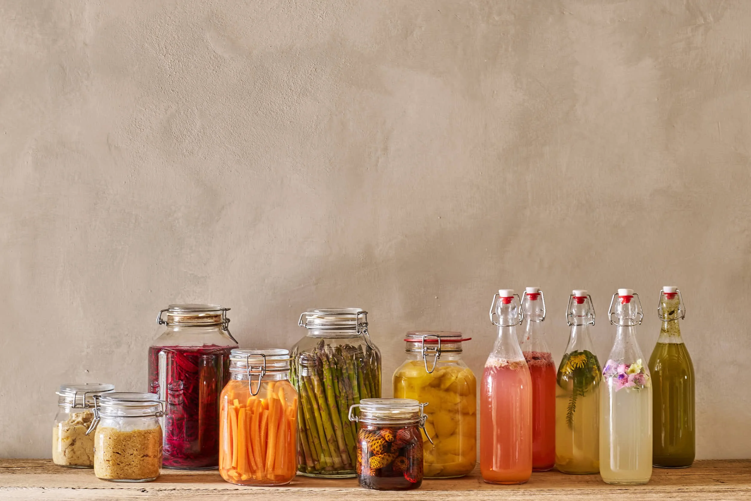 Verschiedene eingelegte Lebensmittel in Gläsern auf einem Holztisch gegen eine beige Wand.