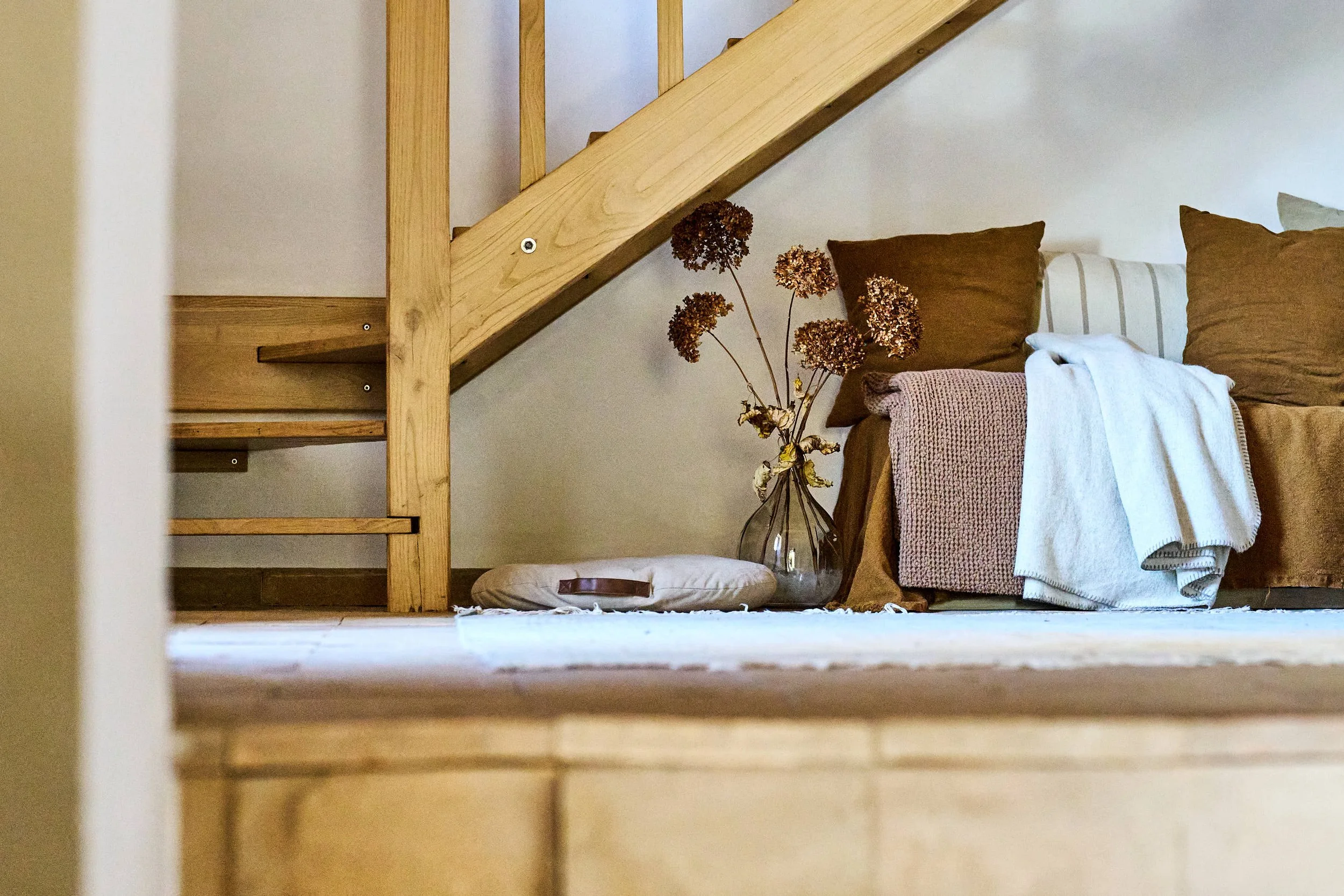 Ein Wohnzimmerbereich unter einer Holztreppe mit einem braunen Sofa, einem weißen Kissen, einem pinkfarbenen Wäschenetz, einem weißen Handtuch, einer Glasvase mit getrockneten Blumen und einem runden Kissen auf einem Teppich.