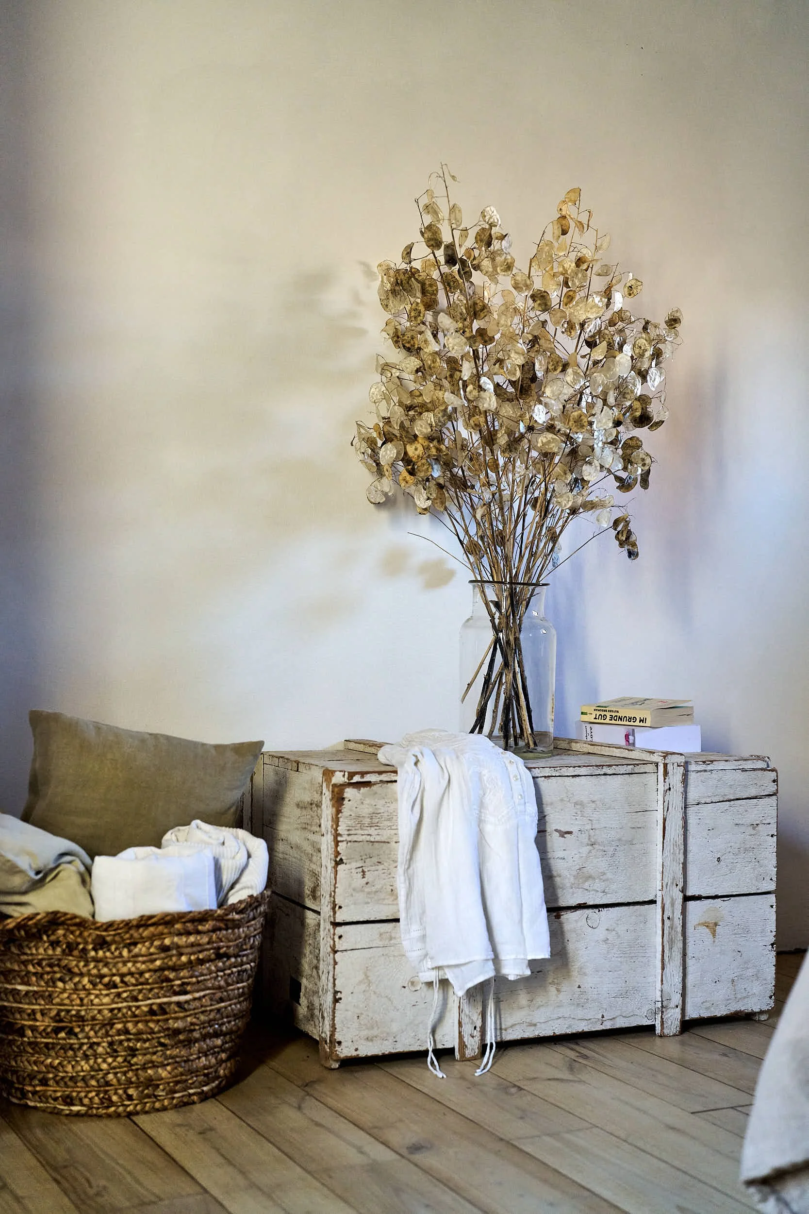 Ein weißer, rustikaler Holztresen mit einer Vase voller Trockenblumen und zwei Büchern darauf. Daneben eine Korbtasche mit weißen Kleidungstücken. An der Wand hängt ein grauer Kissen.
