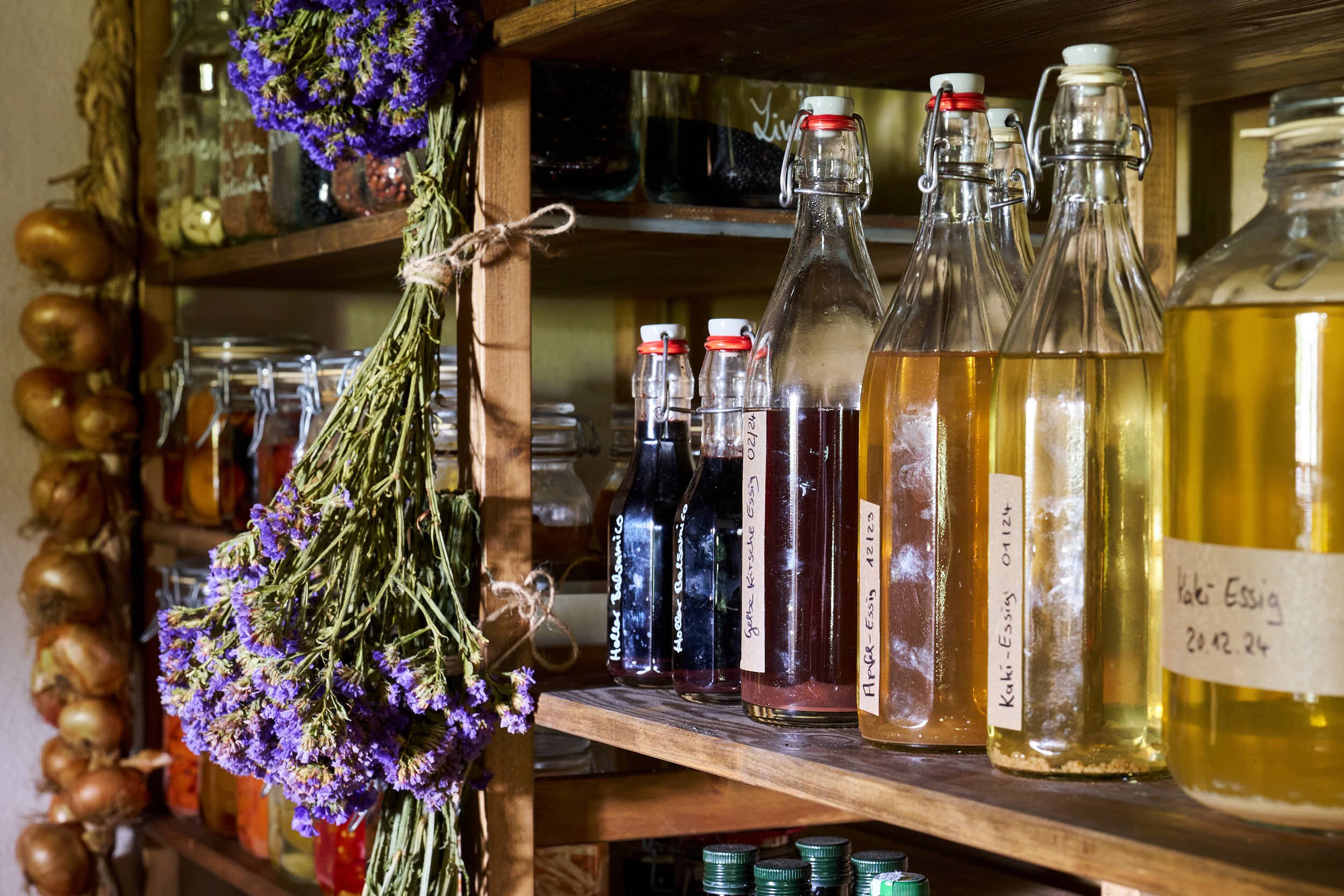 In einem Holzregal stehen Flaschen mit selbstgemachten Essigen und anderen Flüssigkeiten, neben einer getrockneten Lavendelblume und Gläsern.
