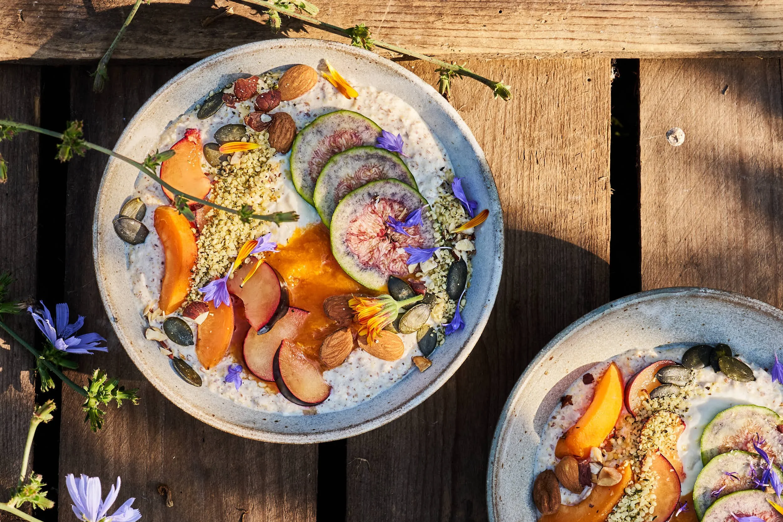 Zwei Schalen mit Joghurt, frischen Pfirsichen, Feigen, Nüssen, Samen und blauen Blüten auf einem Holztisch im Freien.