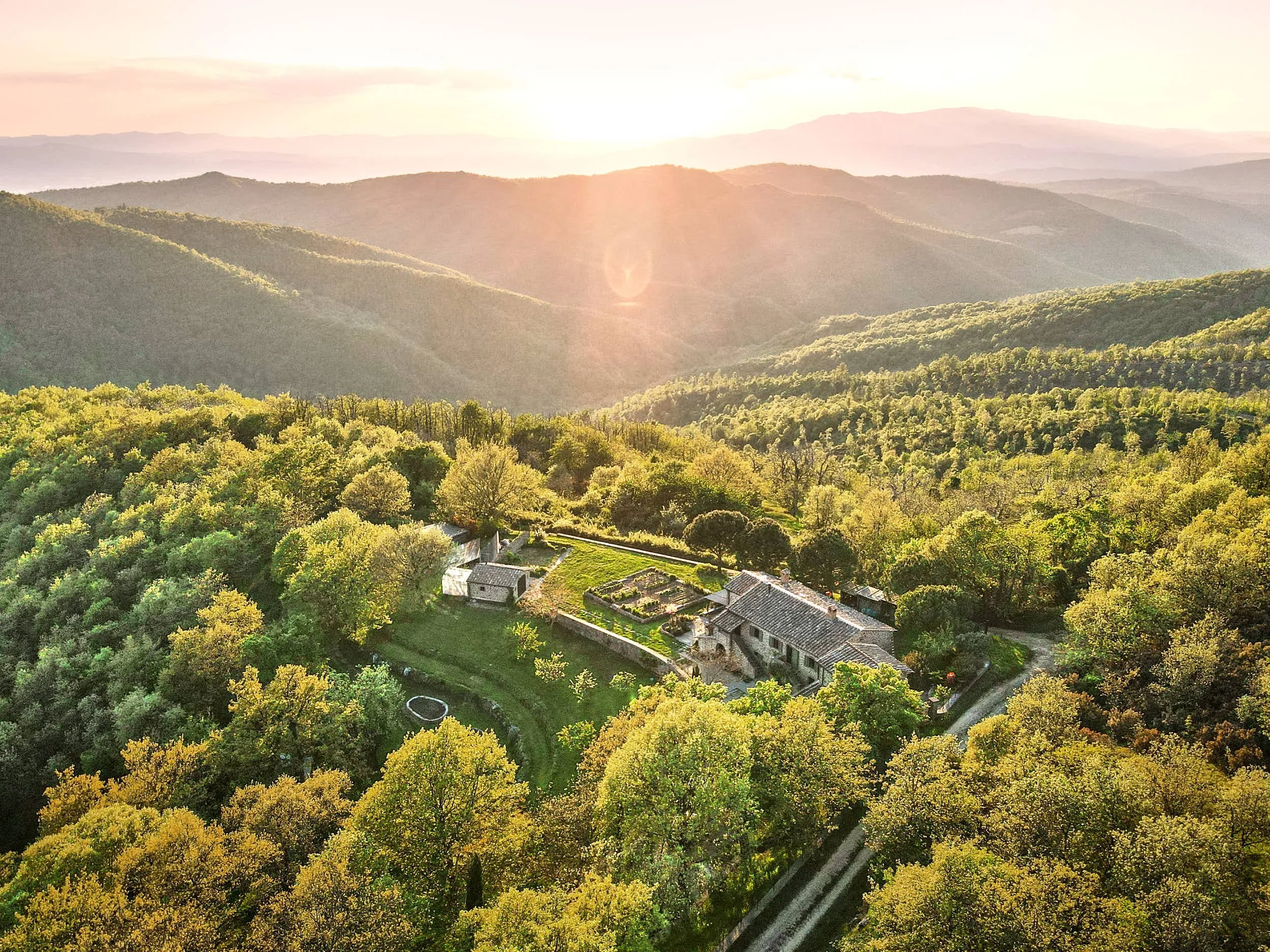 Landschaft mit Bergen, Sonnenuntergang, bewaldetem Gebiet und einem Haus mit Garten in der Vordergrund.