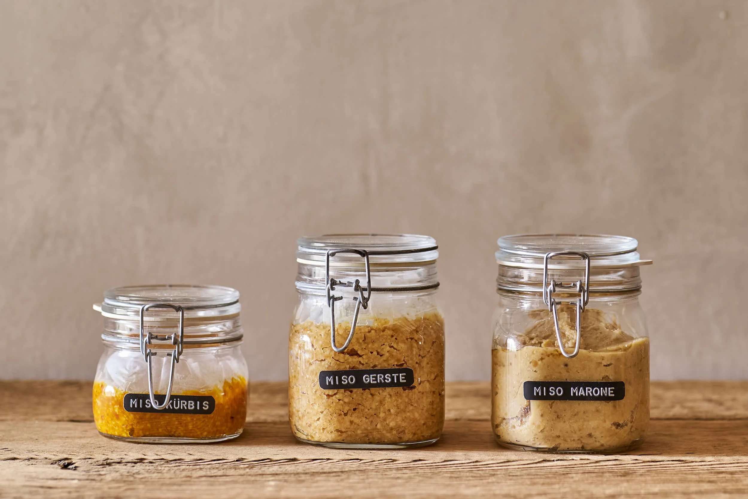 Drei Gläser mit fermentierenden Zutaten auf einem Holztisch, von links nach rechts: Kürbismiso, Gerstemiso, Maoriso, alle in Einmachgläsern mit metallverschluss.