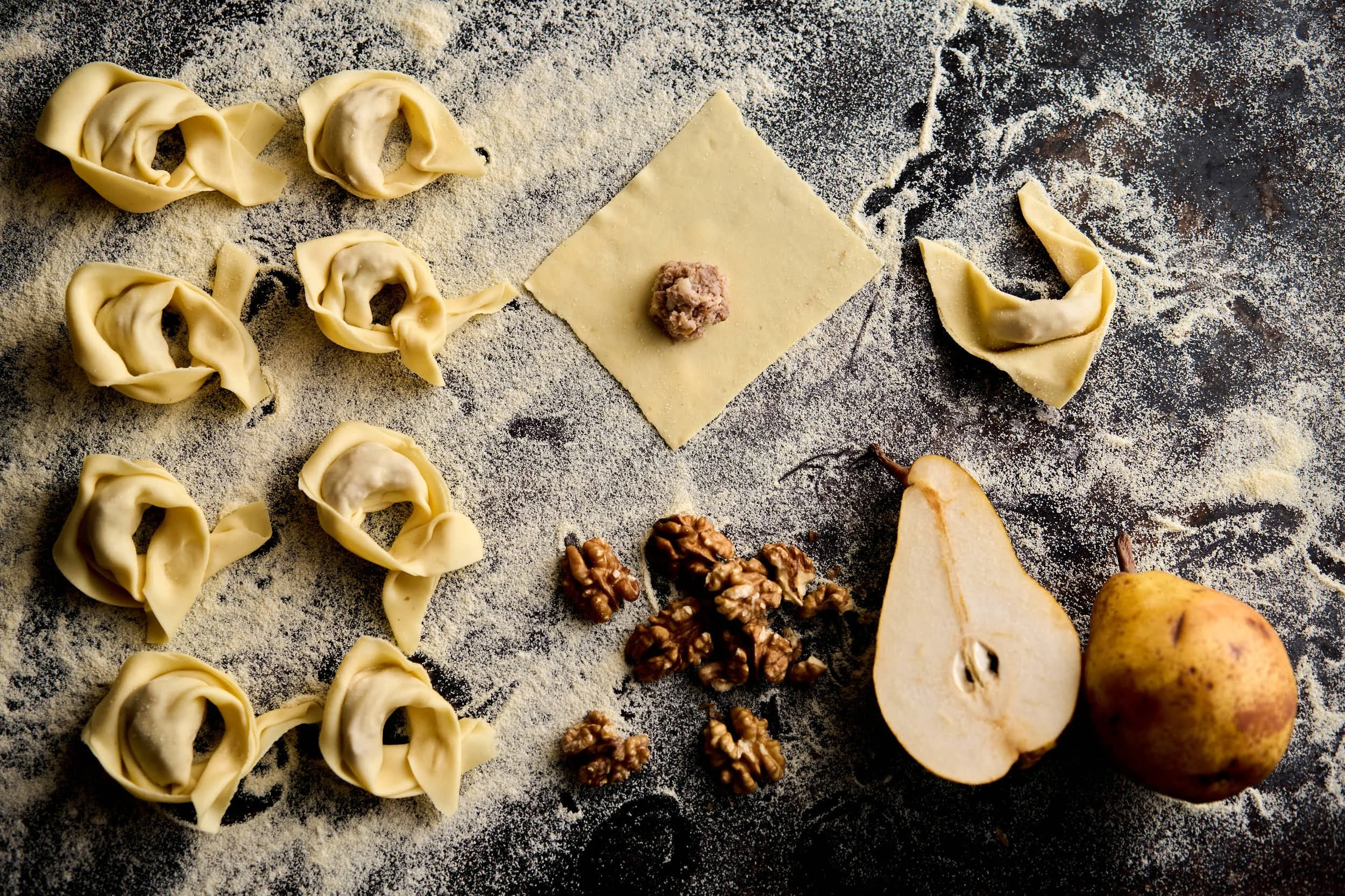 Zubereitung von Teigwaren mit walnüssen, einer Birne, einer halben Birne, einem feststehenden Teigquadrat mit Füllung, alles auf einer mit Mehl bestäubten schwarzen Oberfläche.