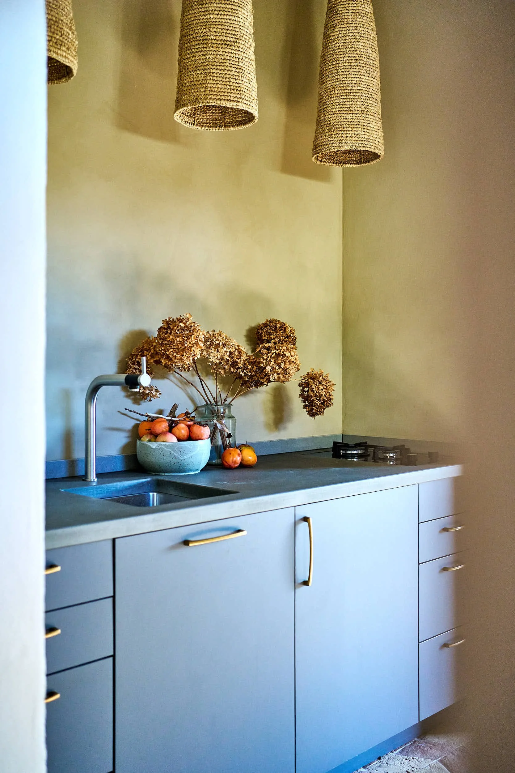 Moderne Küchenschrank mit grauen Türen, eine Spüle, einen Herd, dekorative Trockenblumen in einer Vase, eine Schale mit Äpfeln, hängende Lampen aus Naturmaterialien, gelbe Wände.