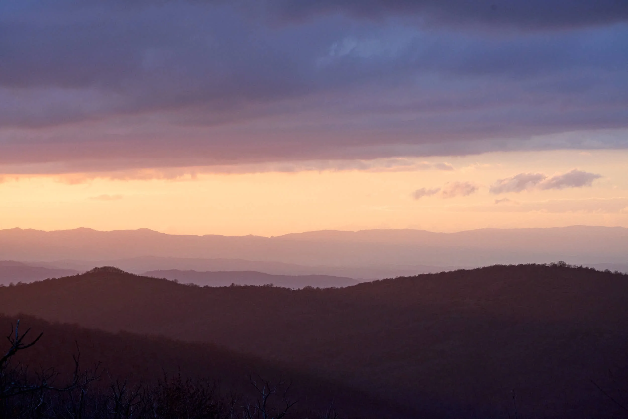 Ansicht eines Bergpanoramas bei Sonnenuntergang mit mehrfachem Hügel- und Bergsilhouette, dunklen Wolken im Himmel und einem sanften Farbverlauf von lila bis orange.