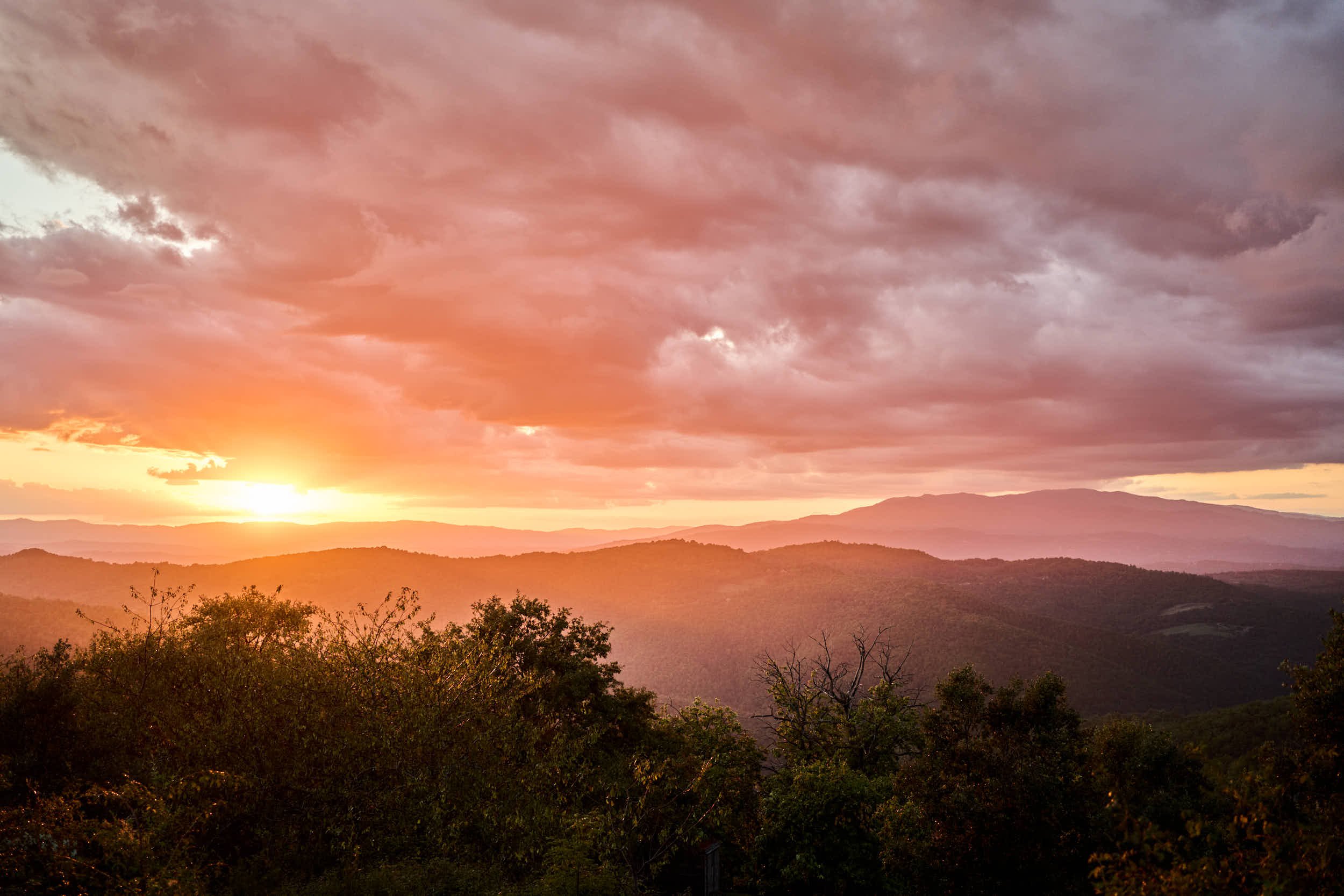 Ein Sonnenuntergang über einer Gebirgs- und Waldlandschaft mit dunklen Wolken am Himmel.