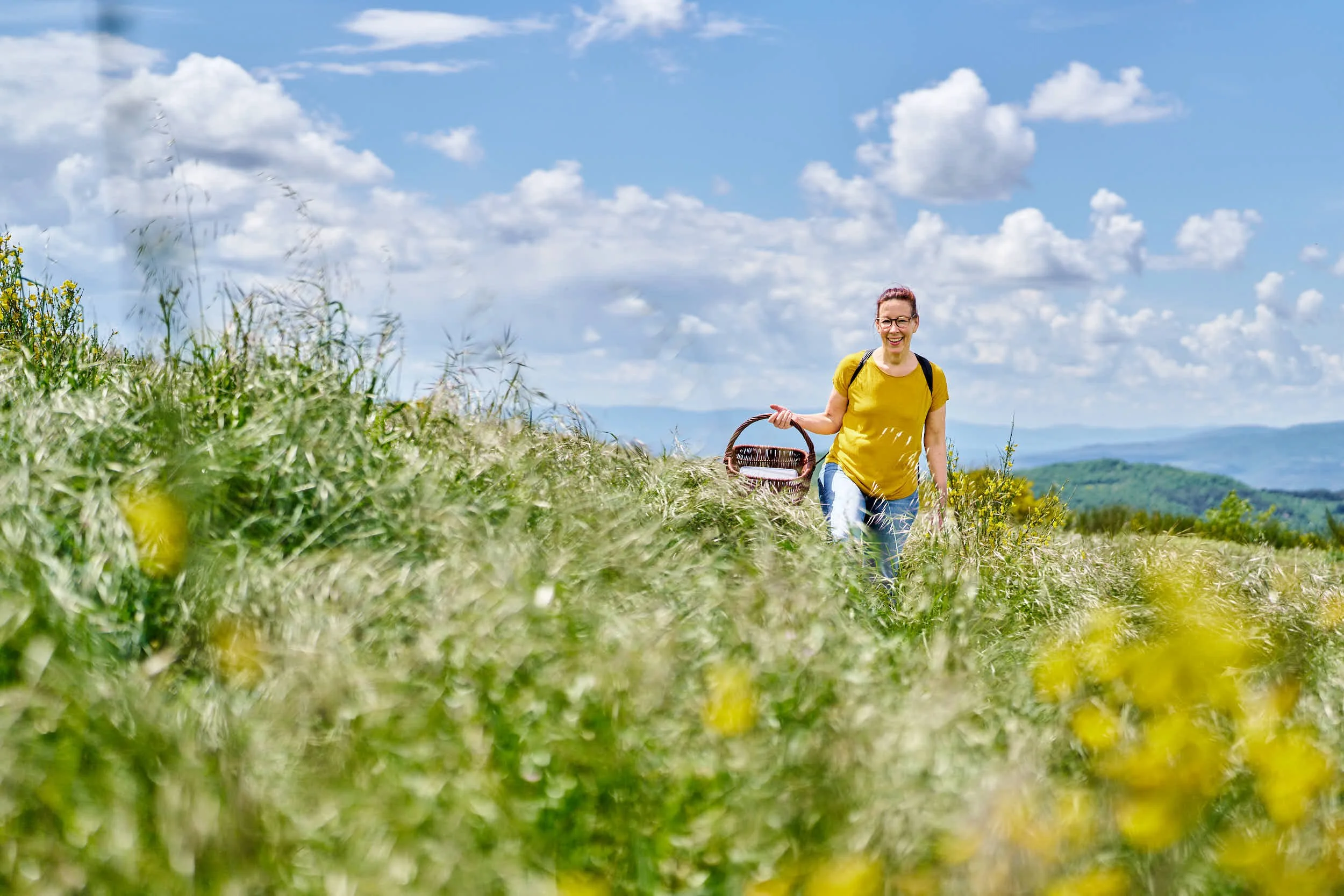 Frau mit Brille trägt gelbes T-Shirt und blaue Jeans, spaziert durch eine grüne Wiese, trägt einen Korb in der Hand und lächelt, im Hintergrund Berge und blauer Himmel mit Wolken.