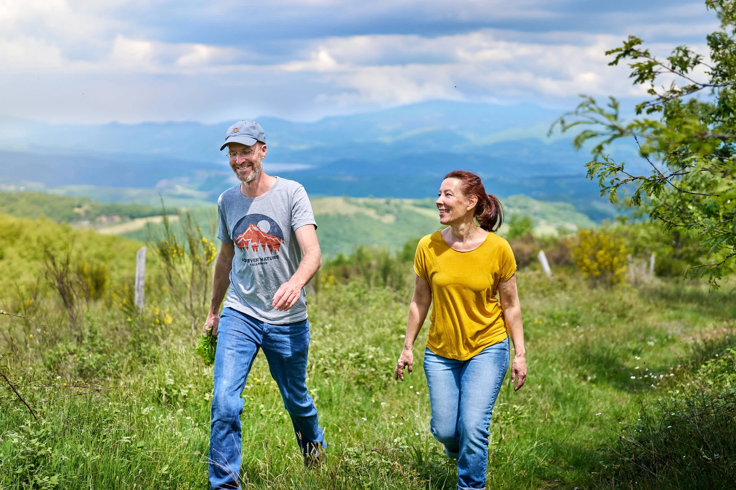 Corinna und Benedikt Fuhrmann spazieren in einer grünen, hügeligen Landschaft mit Bäumen und Wolken am Himmel, lachend und entspannt.