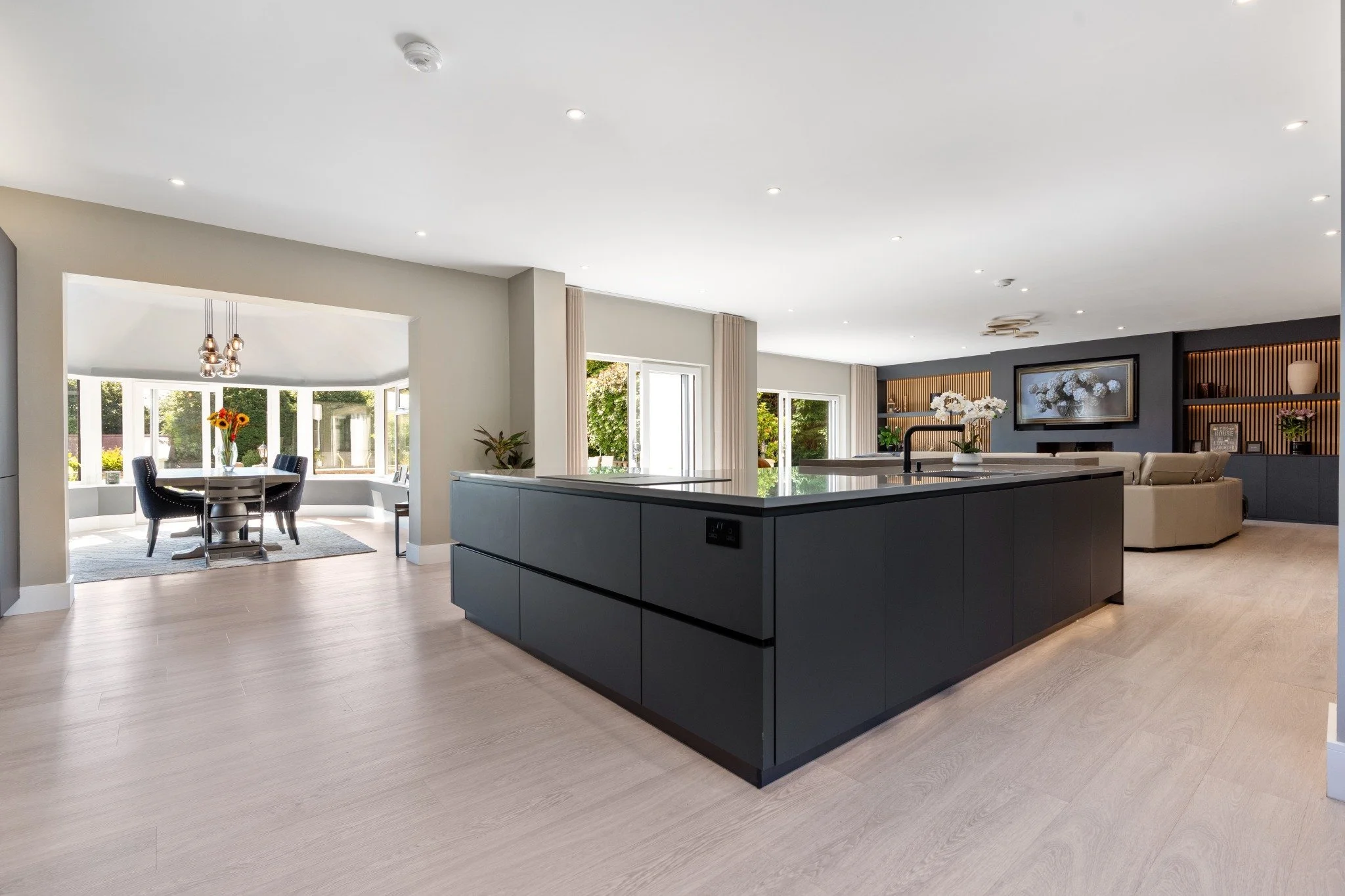 Open-concept living space with a modern kitchen, black island, dining area with a round table and four chairs, and a living room with a wall-mounted TV, beige couches, and built-in shelves with decorative items.