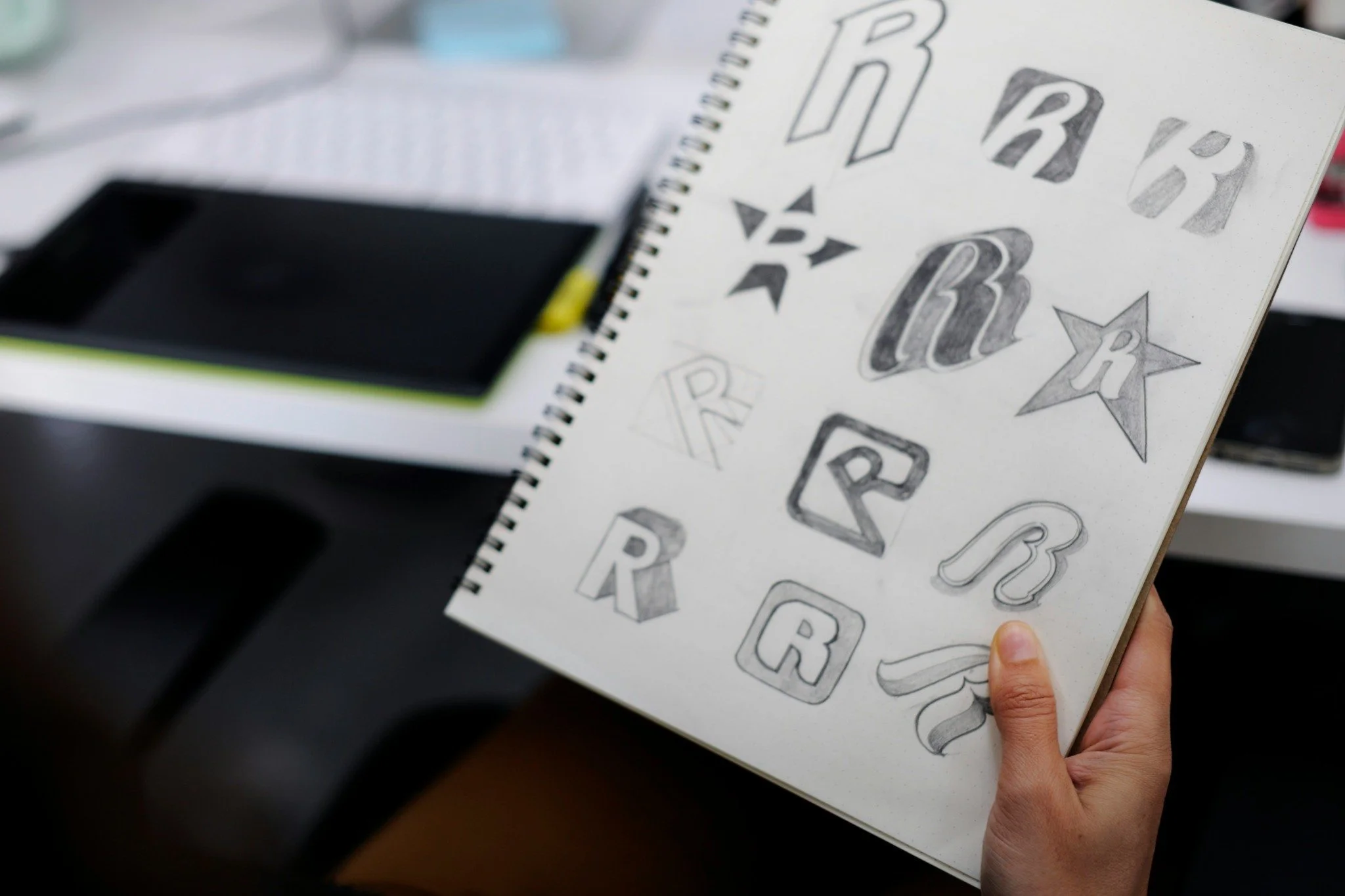 A person holding a sketchbook showing various hand-drawn designs of the letter 'R' in different styles and fonts, with some star-shaped and decorative embellishments.