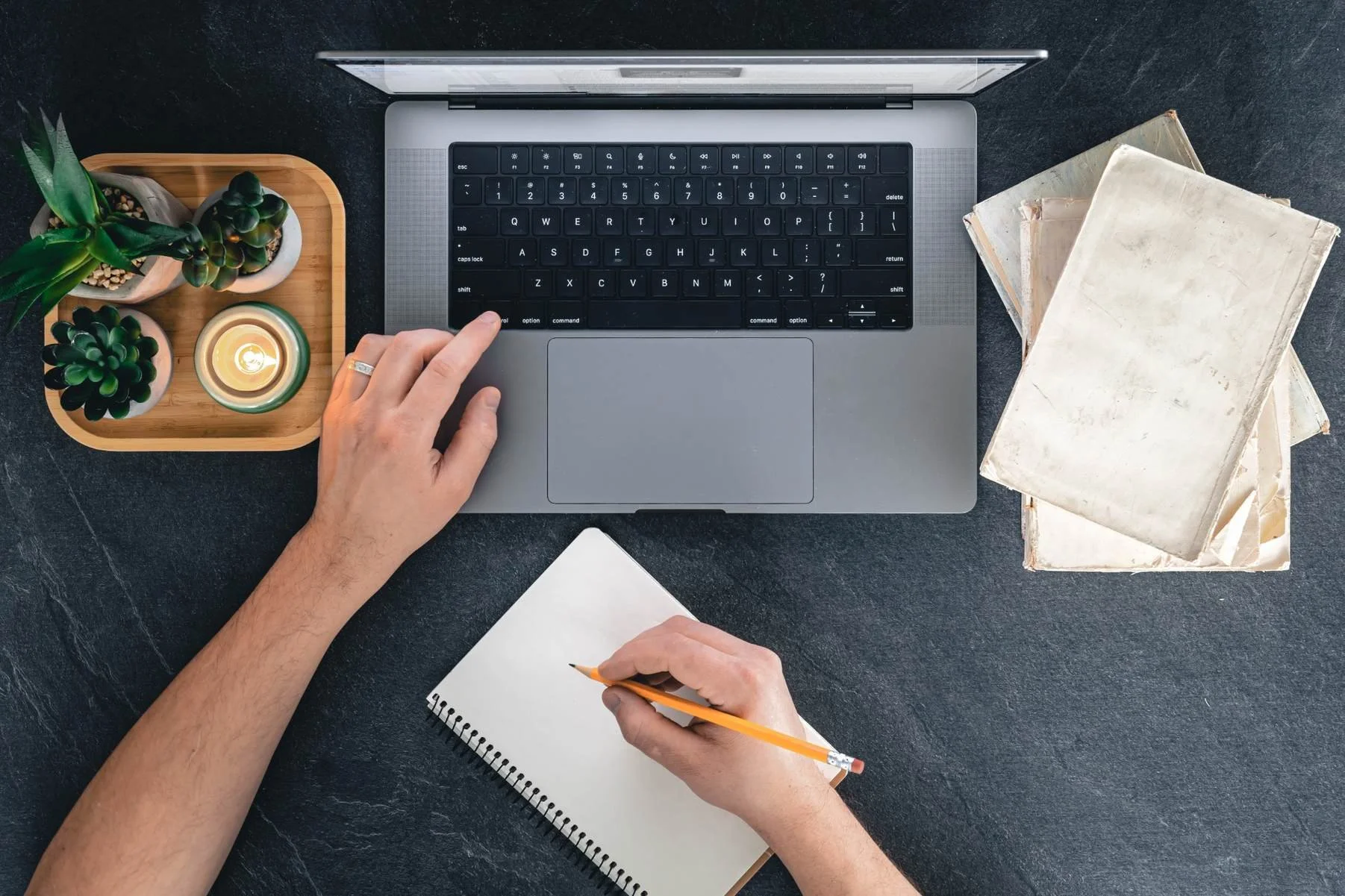 Top view of a workspace with a person using a laptop, taking notes on a notepad, three potted succulents, a candle, and a few old books on a dark surface.