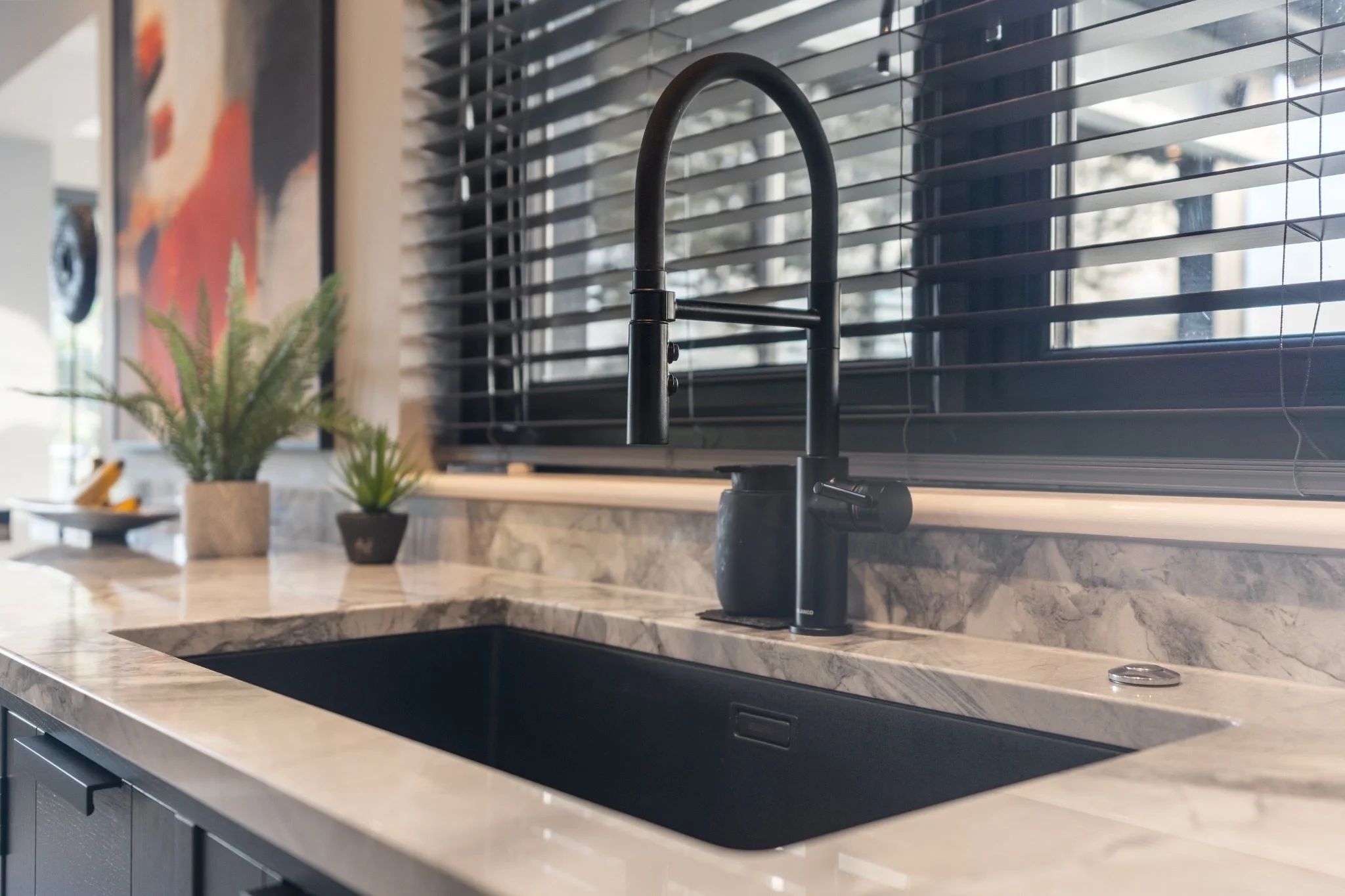 Modern kitchen sink with black faucet on marble countertop near window with blinds, decorative plants, and abstract art on the wall.