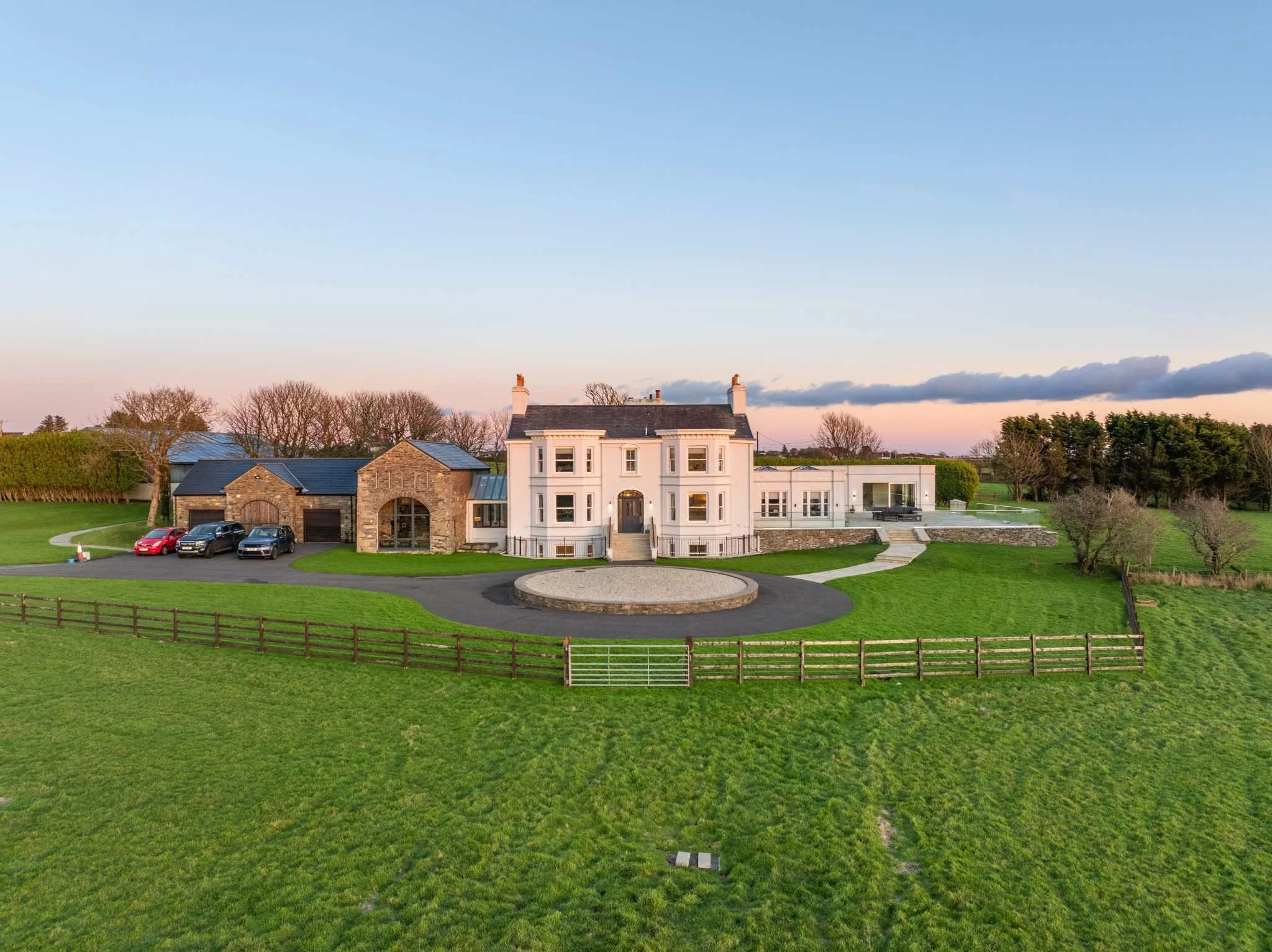 Large white mansion with a circular driveway and a stone fence, surrounded by green grass and trees, under a clear sky at sunset.