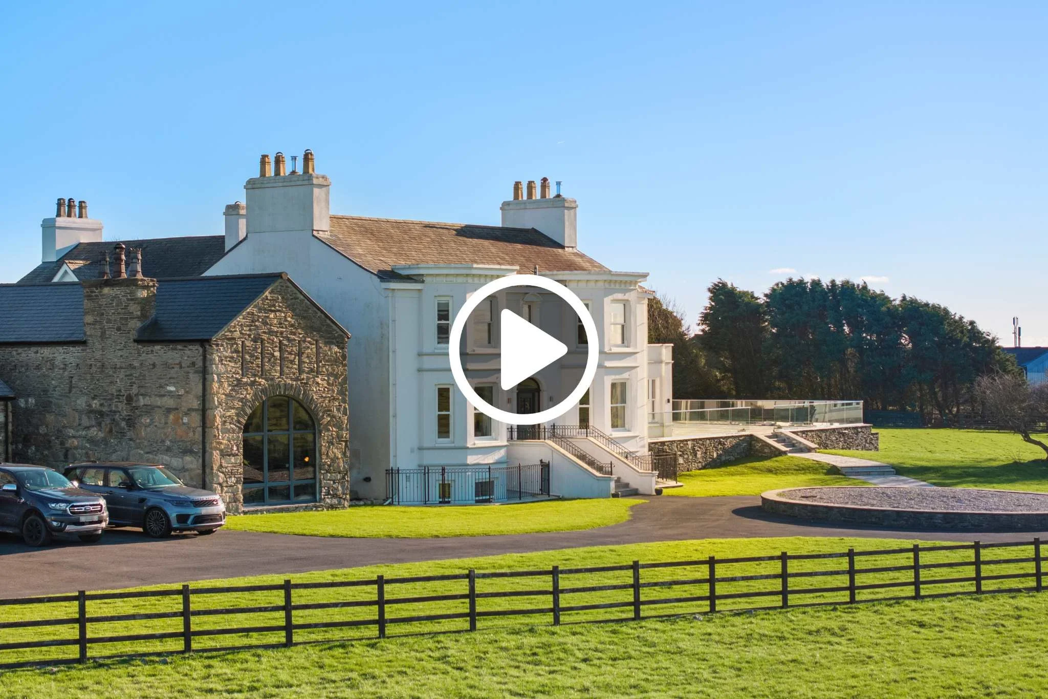 Large white and stone house with a front porch, set on a grassy hill with a fenced yard, parked cars in the driveway, trees in the background, and a clear blue sky.