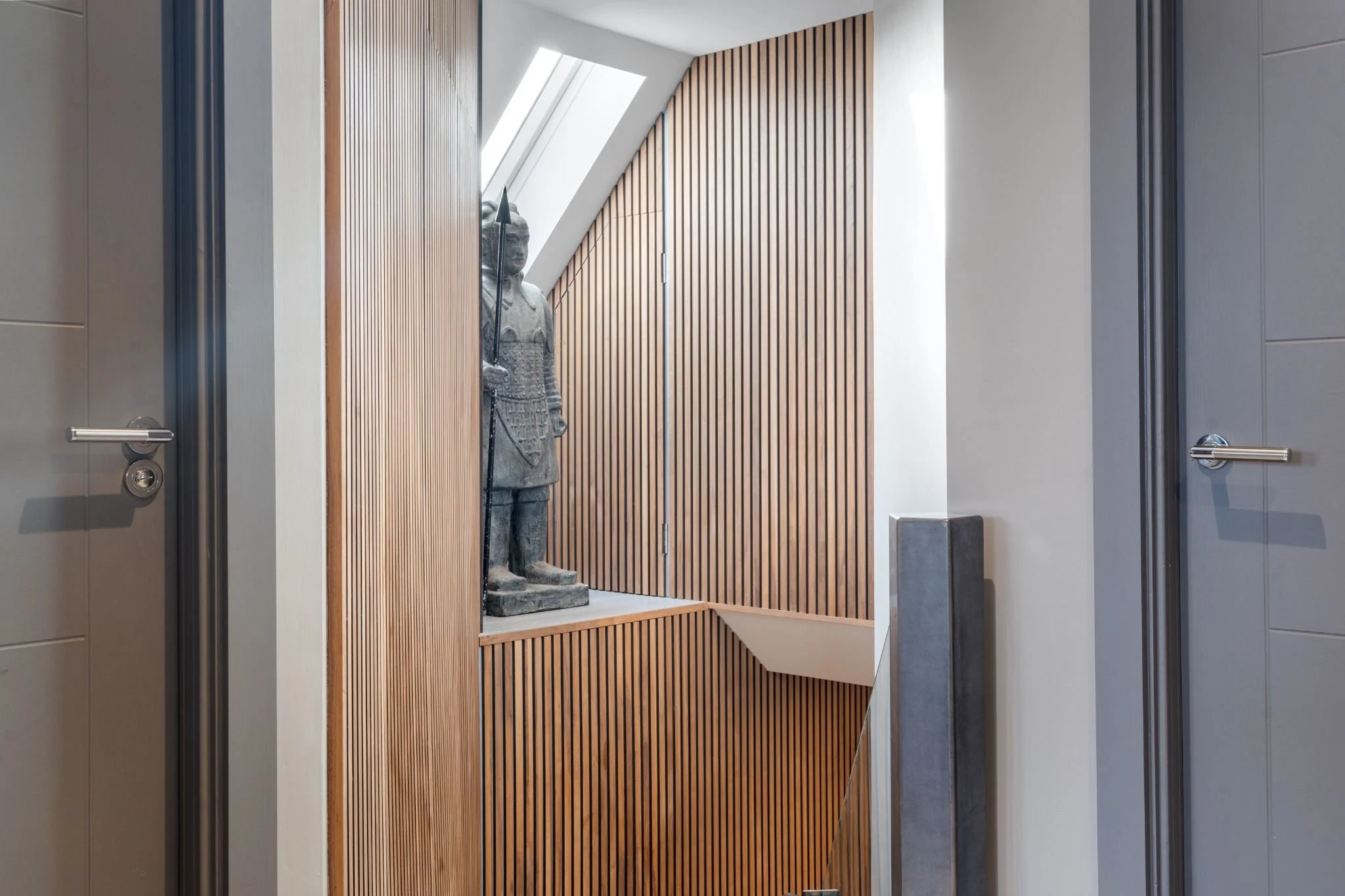 A wooden display area with a stone warrior sculpture holding a spear, located between two gray elevator doors in a modern building interior.