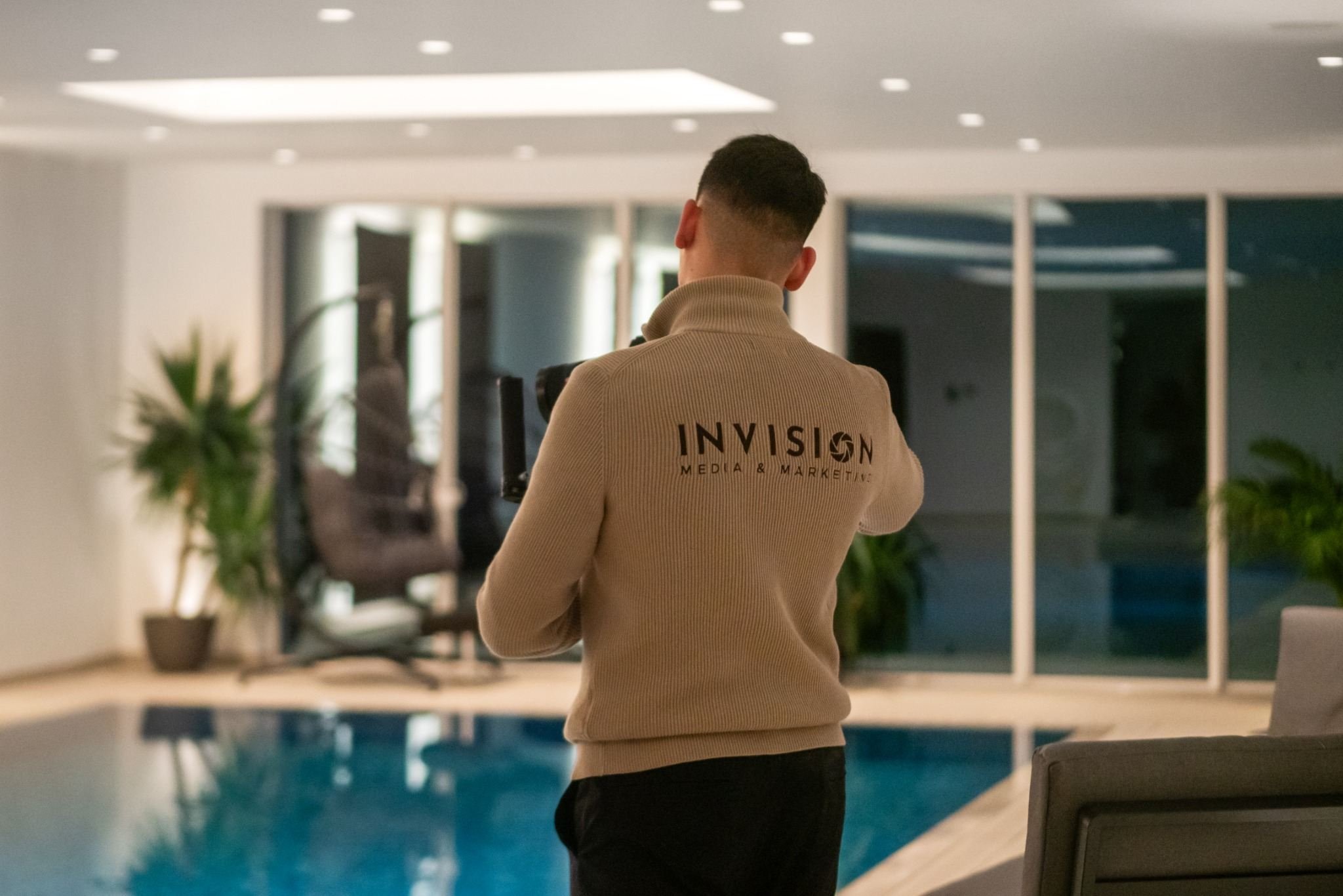 A person standing indoors near a swimming pool, wearing a beige sweater with 'INVISION MEDIA & MARKETING' printed on the back, looking at a camera mounted on a tripod.