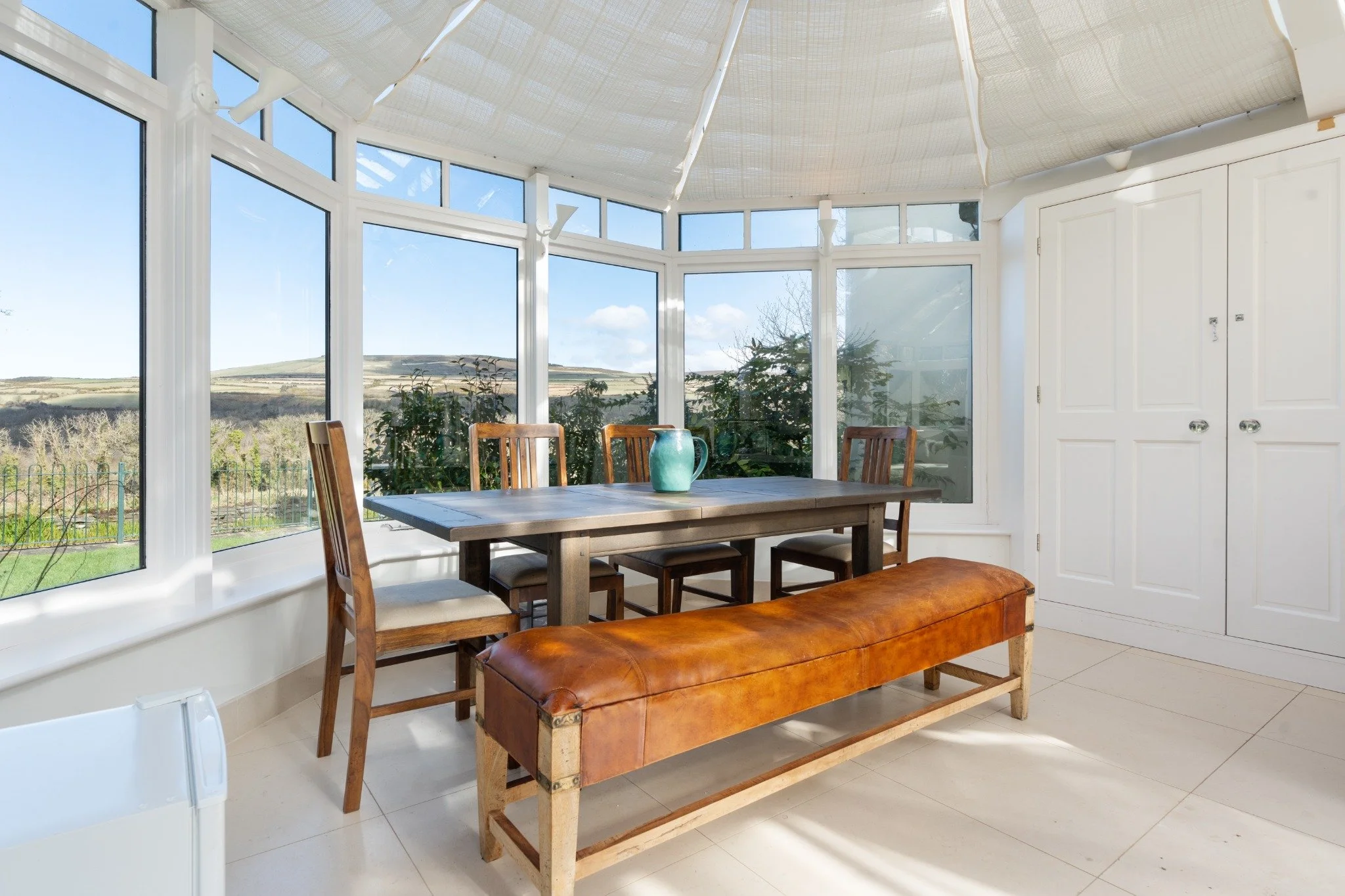 Sunlit dining area with a wooden table, six chairs, a leather bench, and large windows showing a landscape of hills and trees outside.