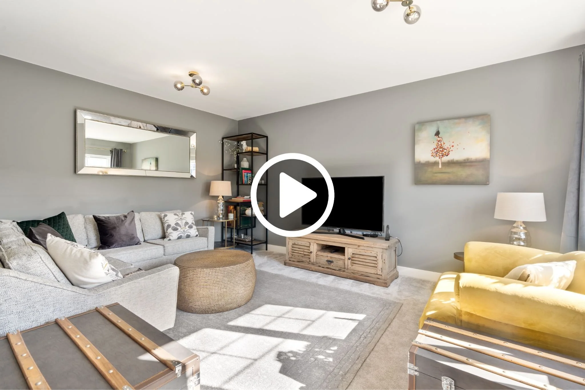 Living room with gray walls, large mirror, gray sofa with throw pillows, yellow armchair, beige ottoman, wooden TV stand with television, bookshelf with decor, and framed artwork on the wall.