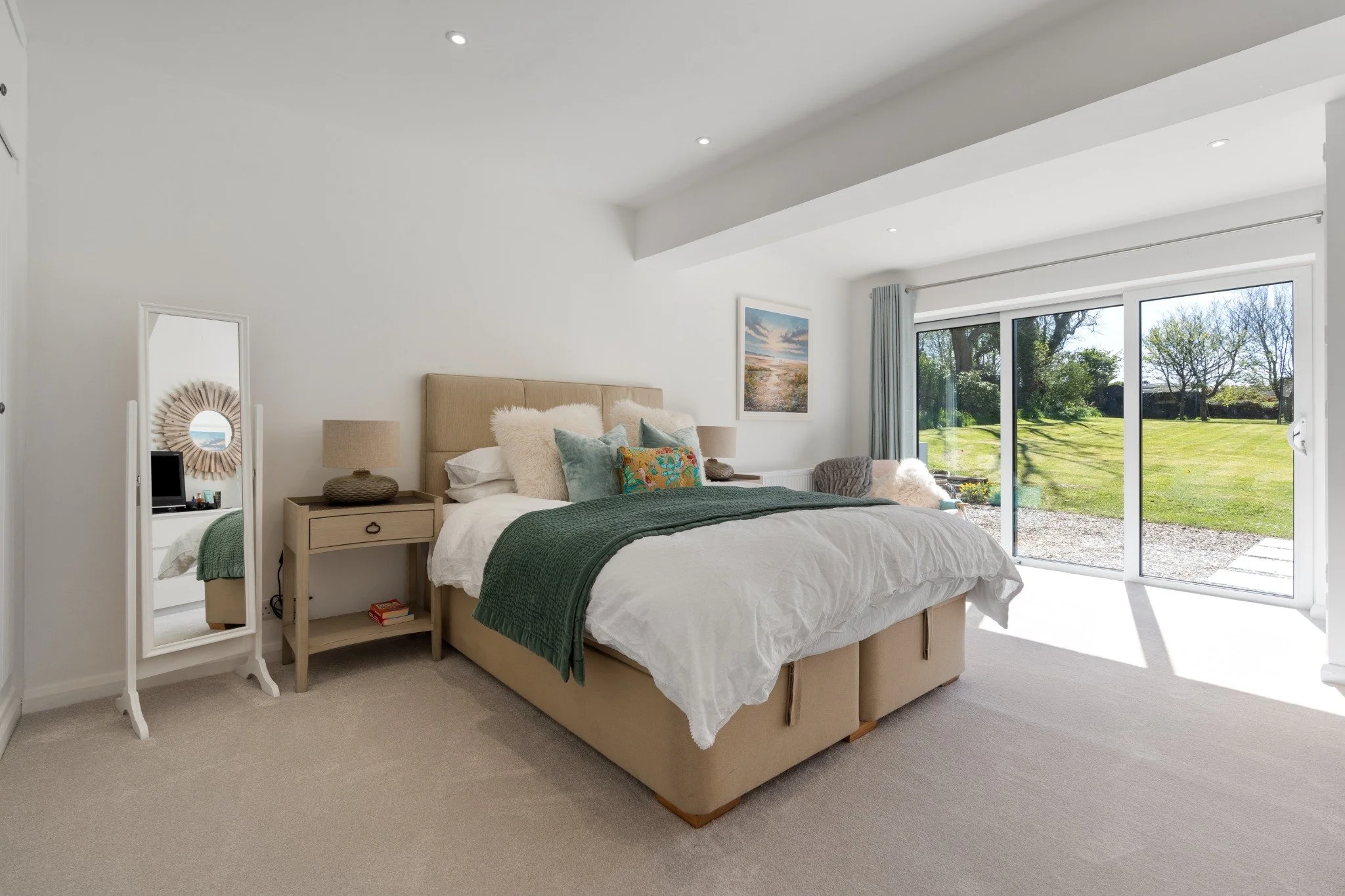 Bright bedroom with large sliding glass doors facing a grassy yard, beige bed with white bedding and decorative pillows, small side table with lamp, and a standing mirror