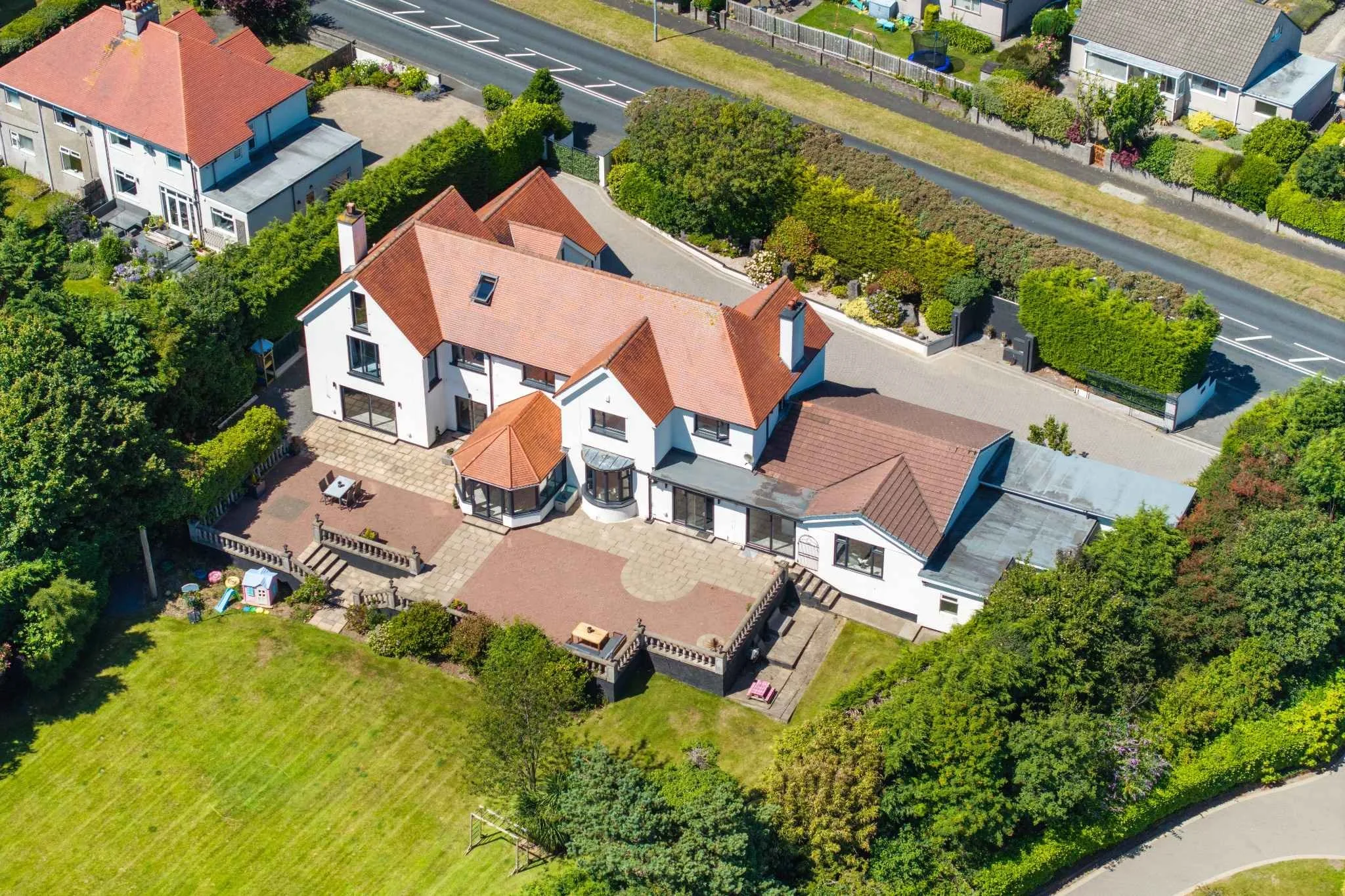 Aerial view of a large white house with a red tiled roof, surrounded by a landscaped yard, garden, and trees, with neighboring houses and a street nearby.
