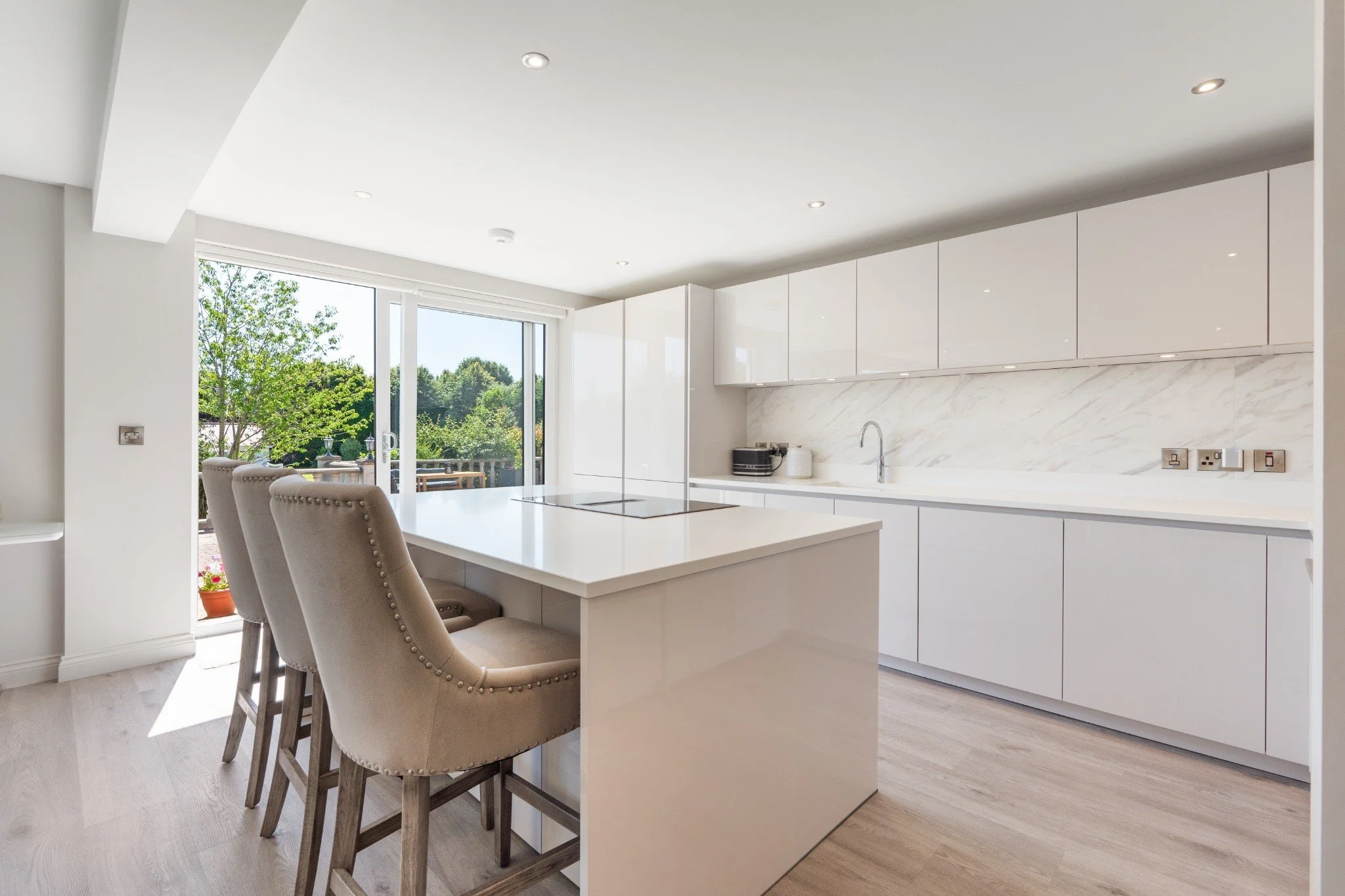 Modern bright kitchen with white cabinets, marble backsplash, and an island with beige upholstered barstools. Large sliding glass doors lead outside to a green backyard.