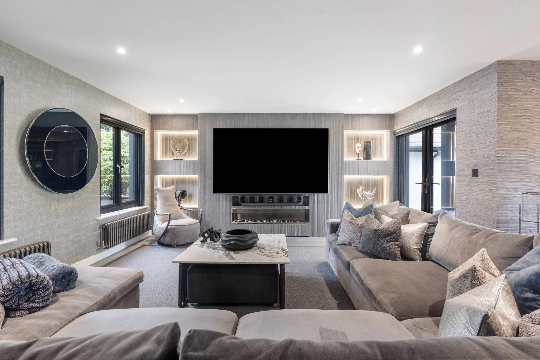 Modern living room with beige sofas, decorative pillows, a marble coffee table, a wall-mounted flat-screen TV, built-in shelves with decor, large windows, and sliding glass doors leading outside.