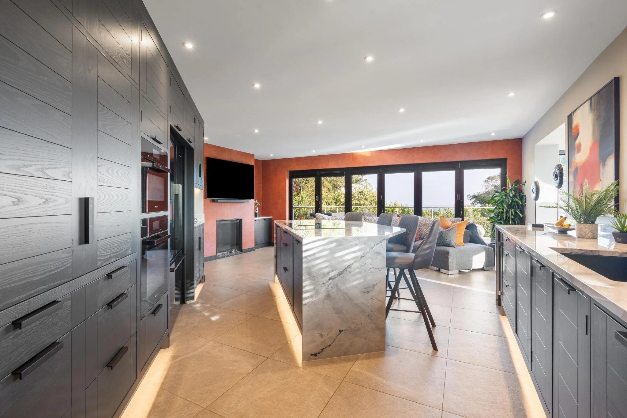 Modern open-concept kitchen and living room with gray cabinetry, a marble island, large windows, and a wall-mounted television.
