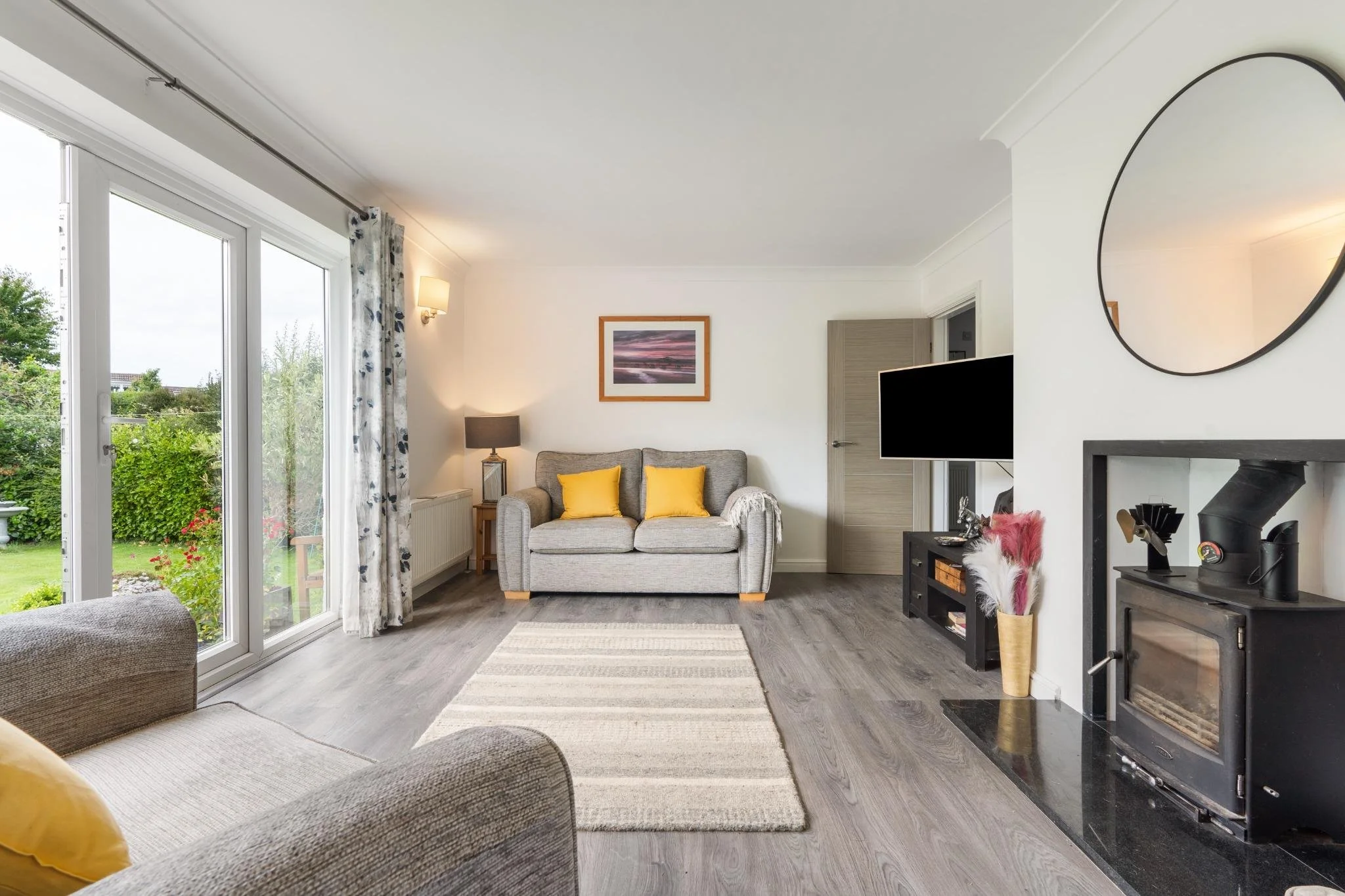 Living room with gray couches, yellow pillows, a window with floral curtains, a flat-screen TV, a fireplace, and decor.