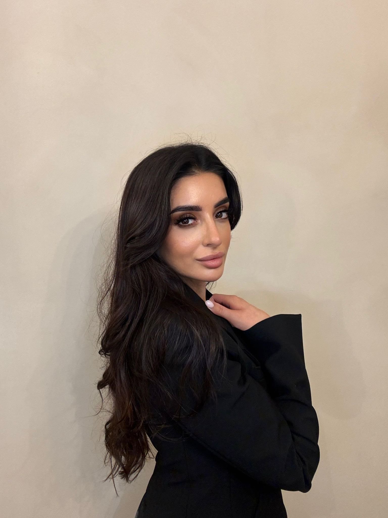 A woman with long, dark wavy hair and makeup posing against a plain beige wall.