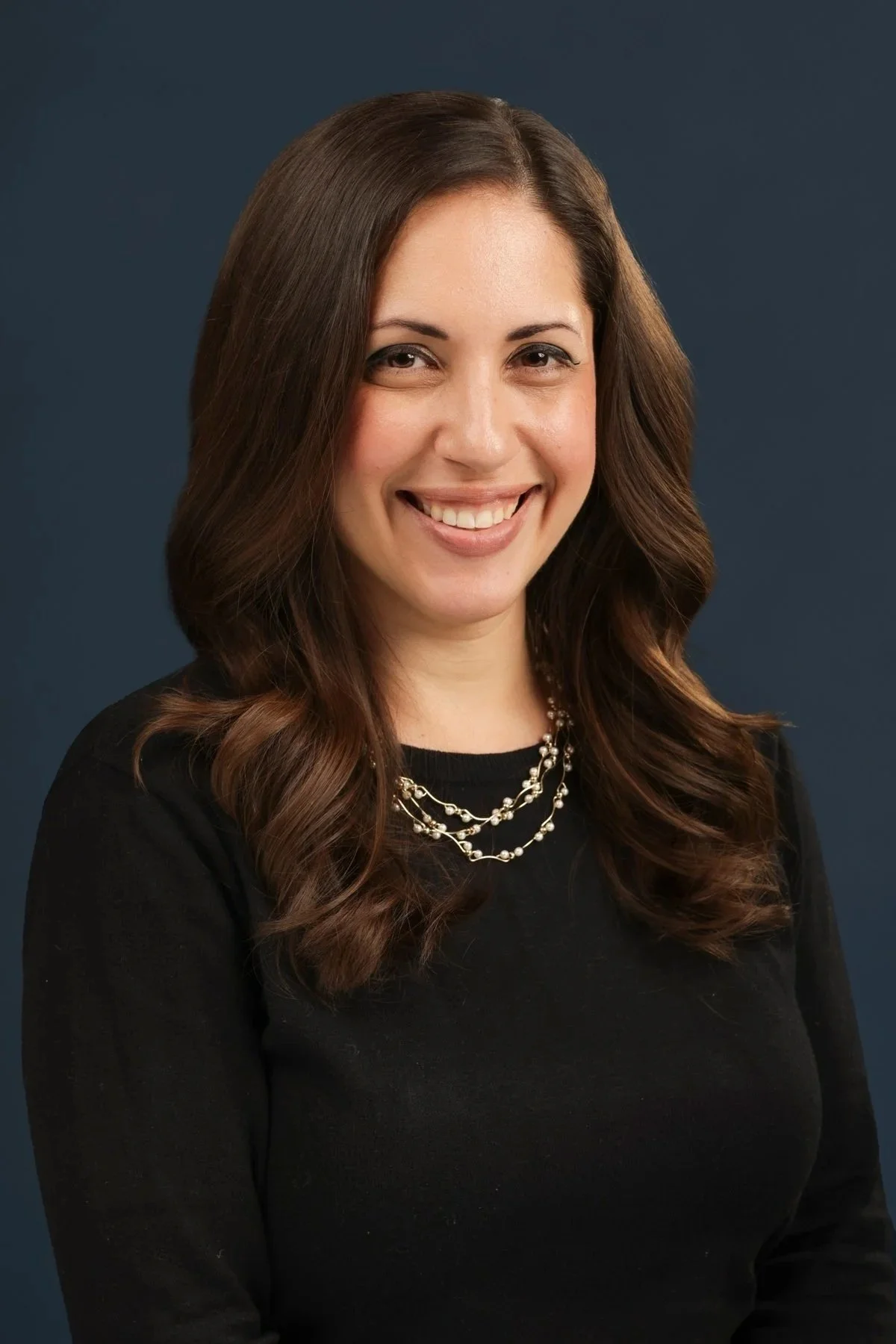 Professional headshot of a woman with brown, wavy hair, wearing a black top and a multi-layered pearl necklace, smiling against a dark blue background.