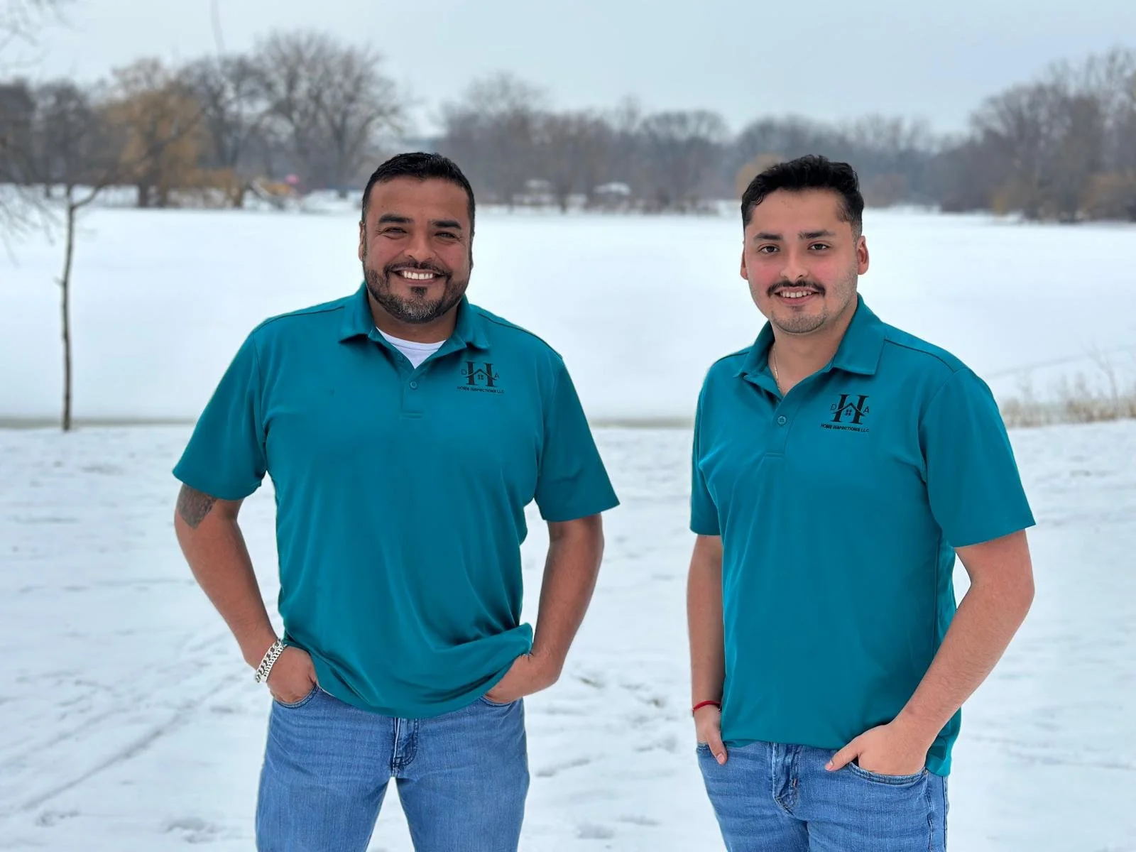 Two trustworthy men standing outdoors in a snowy landscape, both wearing teal polo shirts and jeans, smiling at the camera.