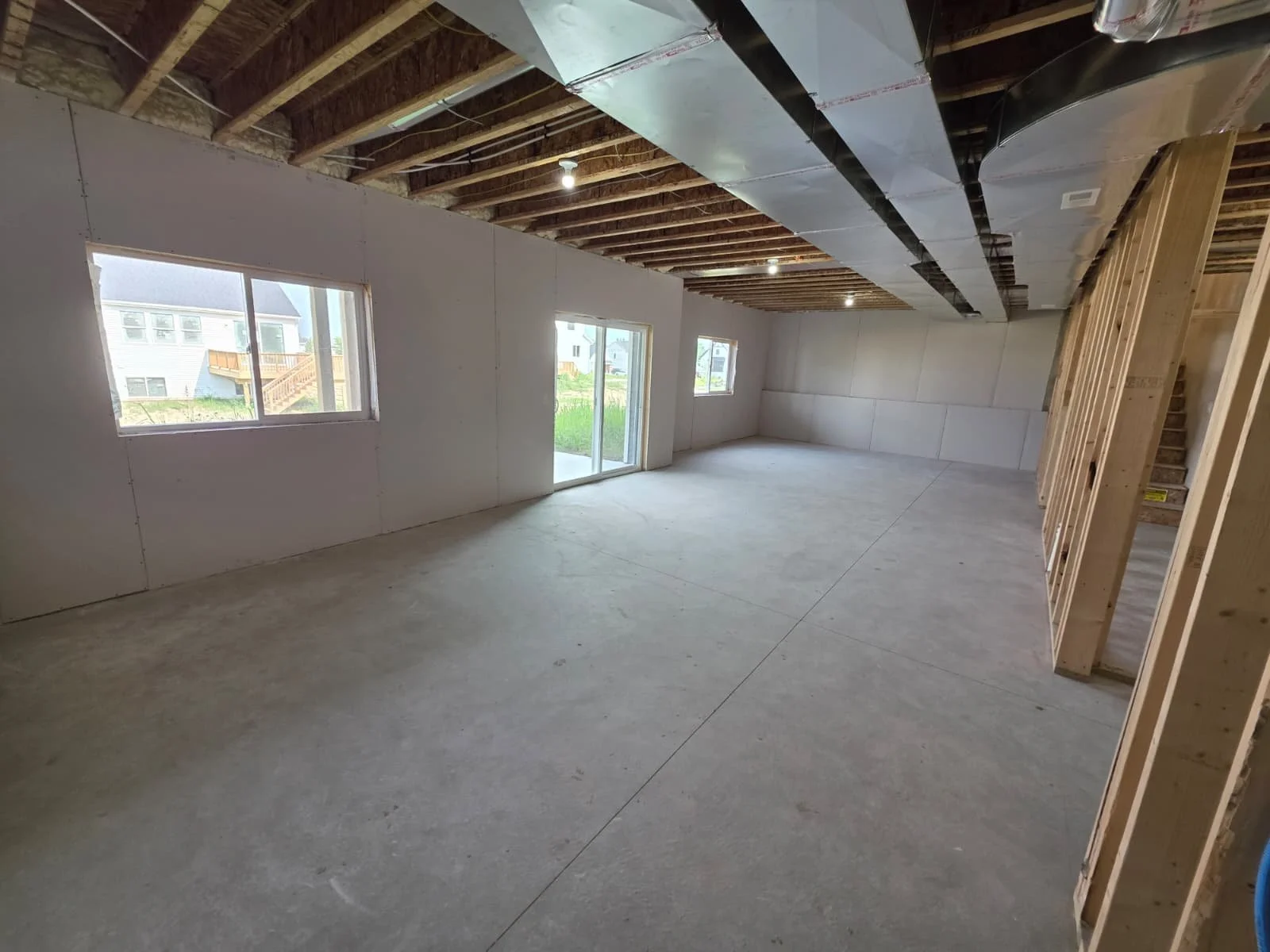 Interior view of an under-construction room, showing drywall on walls, unfinished ceiling with exposed beams and ductwork, and a concrete floor. There are two windows and a sliding door offering outside views.