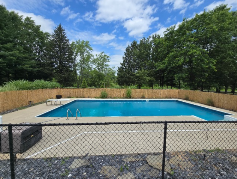An outdoor swimming pool enclosed by a wooden privacy fence and a chain-link fence, with a blue sky and green trees in the background.