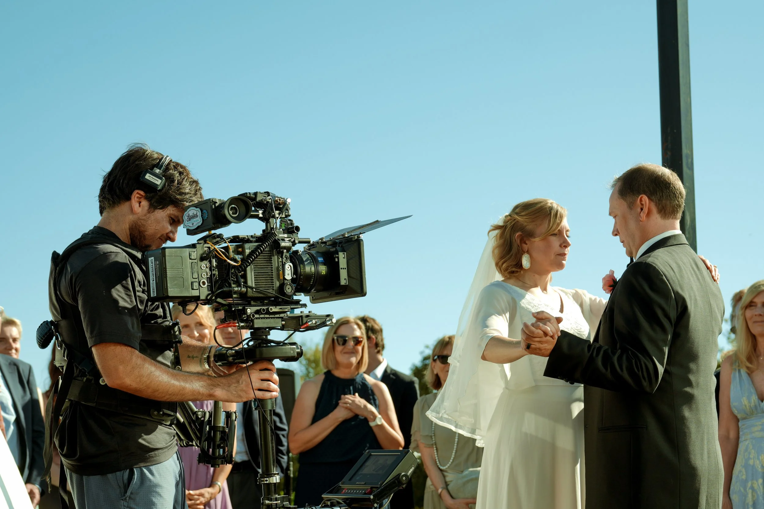 A bride and groom holding hands during their wedding ceremony outdoors, with a professional photographer filming them, and guests watching.