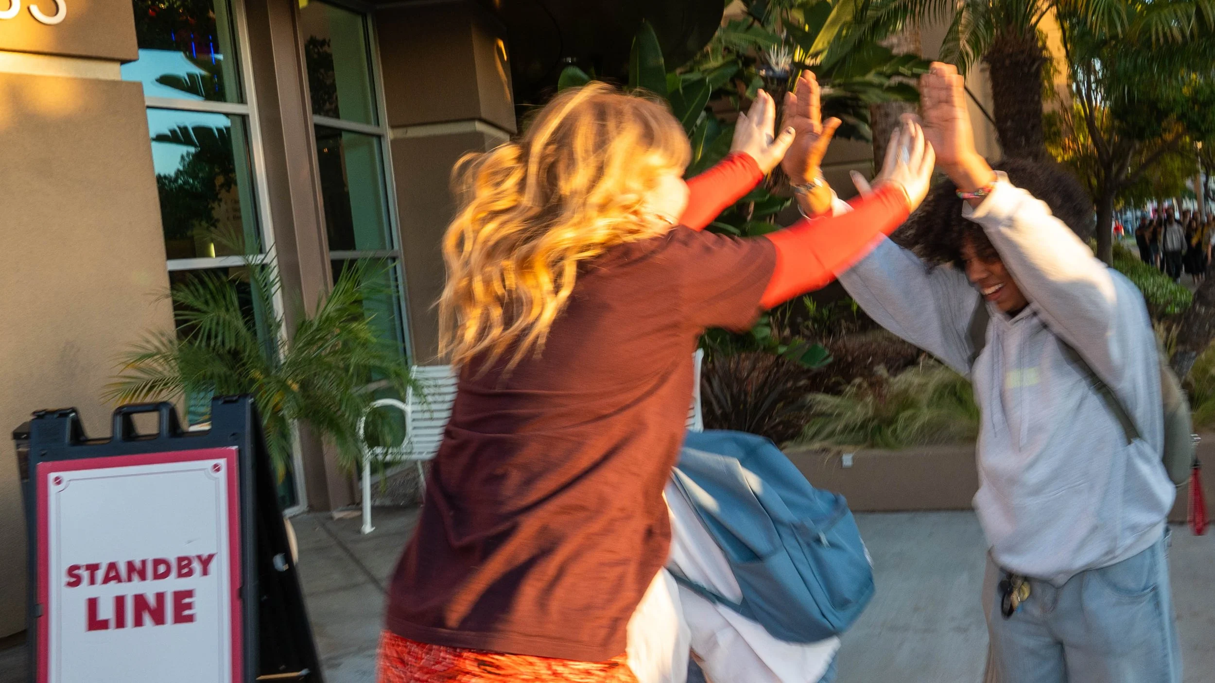 Two women are high-fiving outside a building with glass windows and lush green plants nearby. They are smiling and appear to be celebrating or greeting each other.