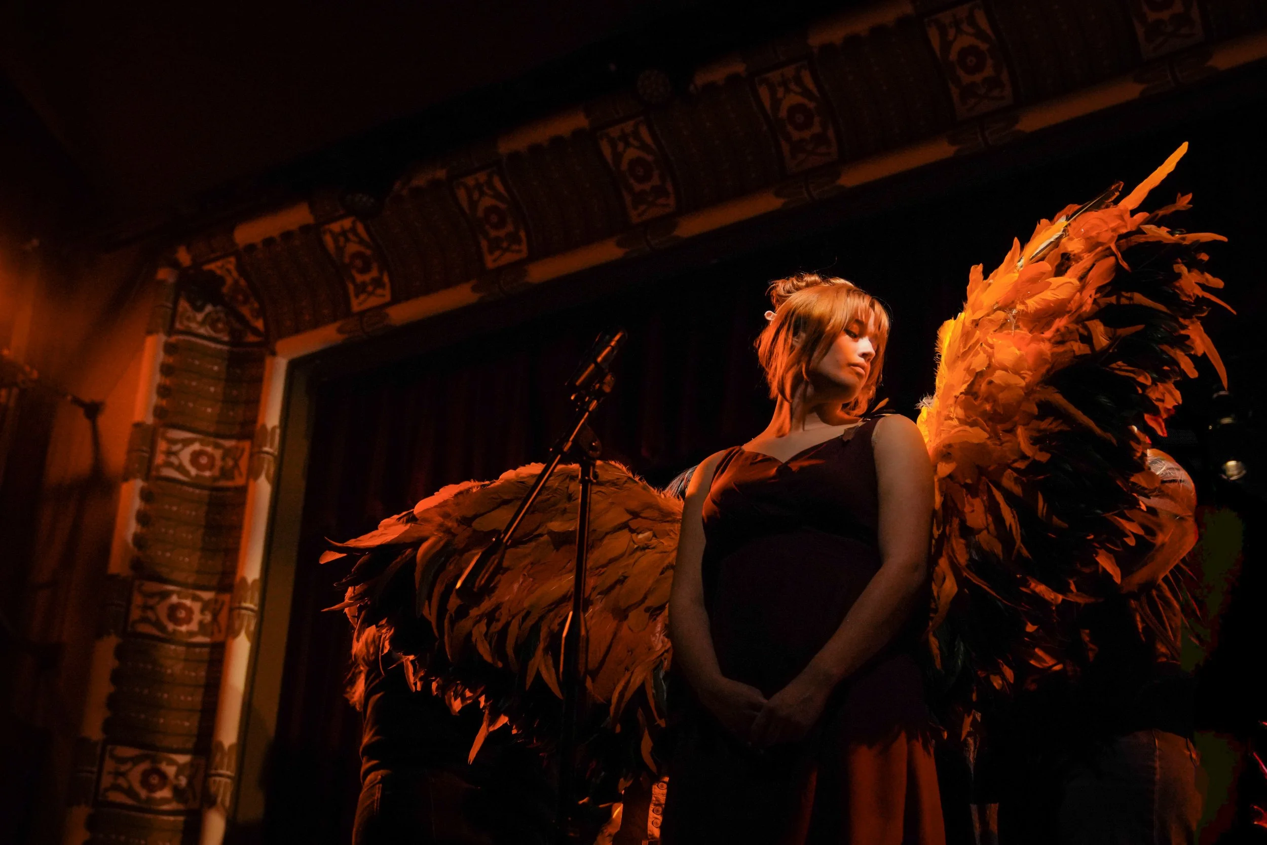 A woman with short hair dressed in a black top standing on stage with large yellow-orange feathered wings, in a dimly lit venue with decorations and curtains.