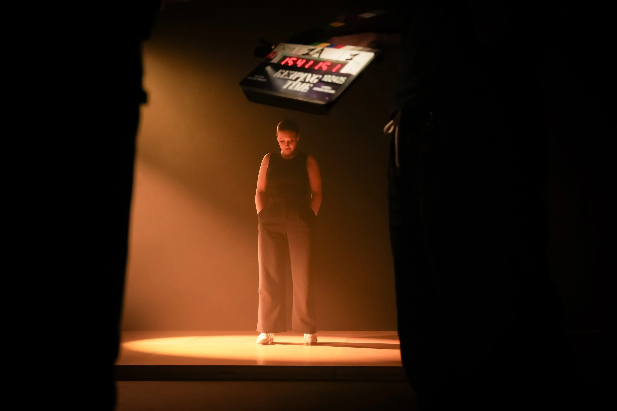 A person standing on stage in spotlight, wearing wide-legged trousers and a sleeveless top, with a blurred background containing a sign or screen.