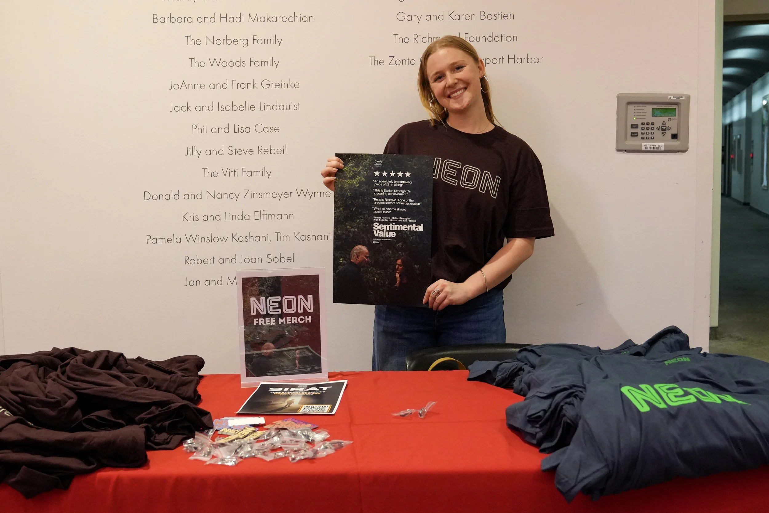 A young woman with red hair and hoop earrings, smiling, holding a magazine at a table with promotional merchandise, including T-shirts, pins, and a flyer, in front of a wall with text listing names and organizations.