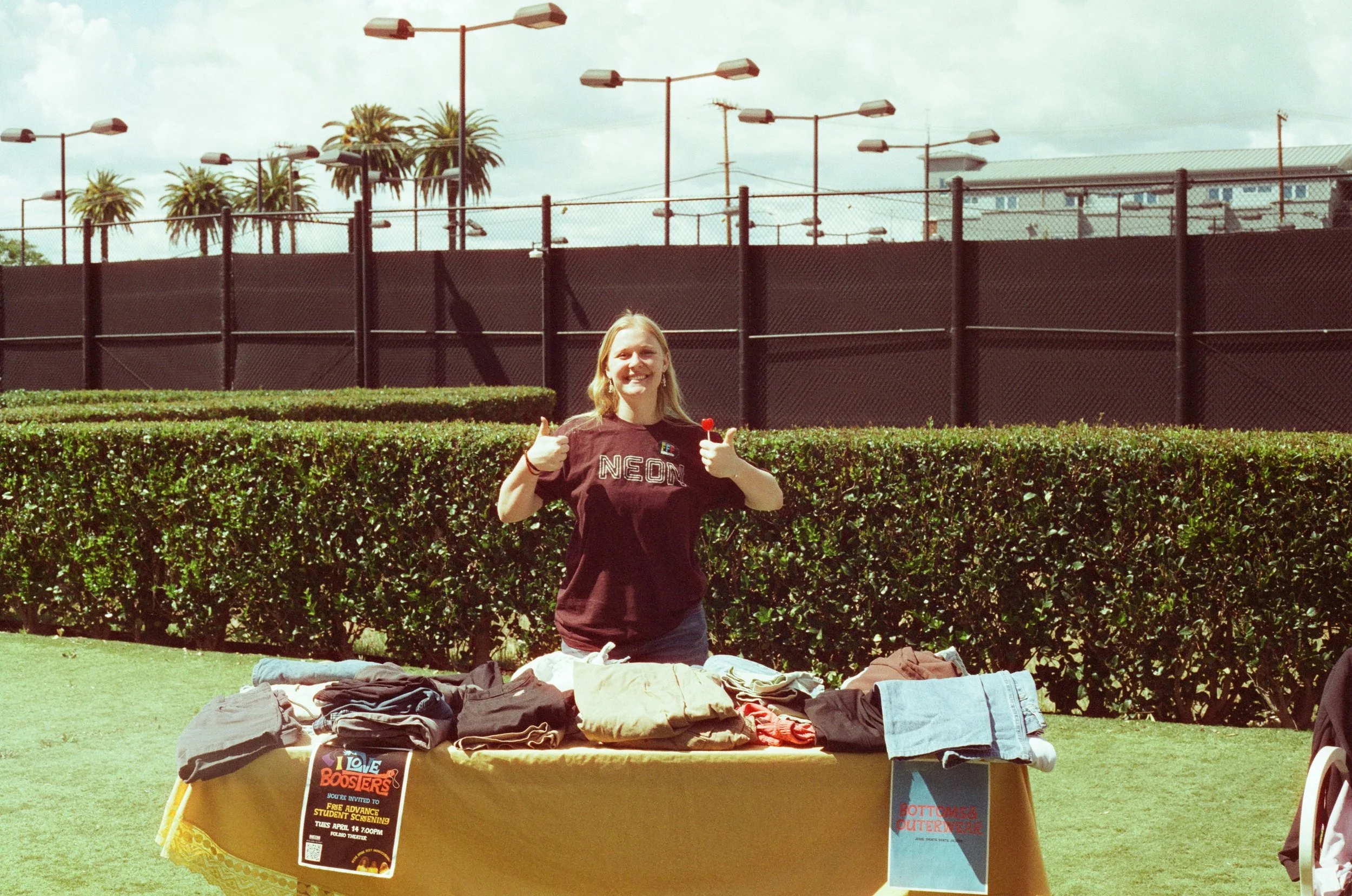 A young woman smiling and giving a thumbs-up behind a table of clothes at an outdoor yard sale with palm trees and a chain-link fence in the background.