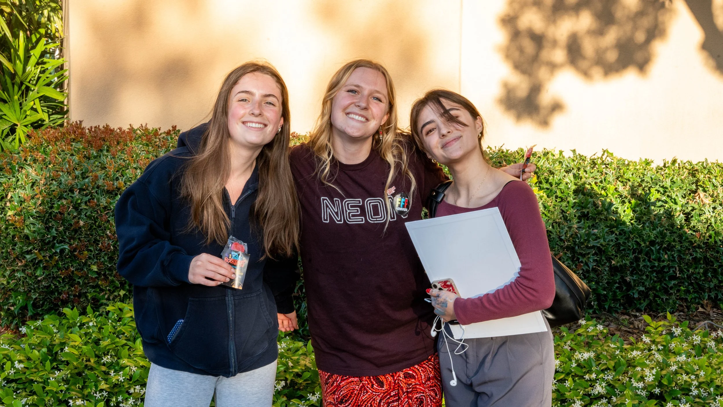 Three young women smiling and standing outdoors near bushes with green leaves and small white flowers, with a beige wall in the background.