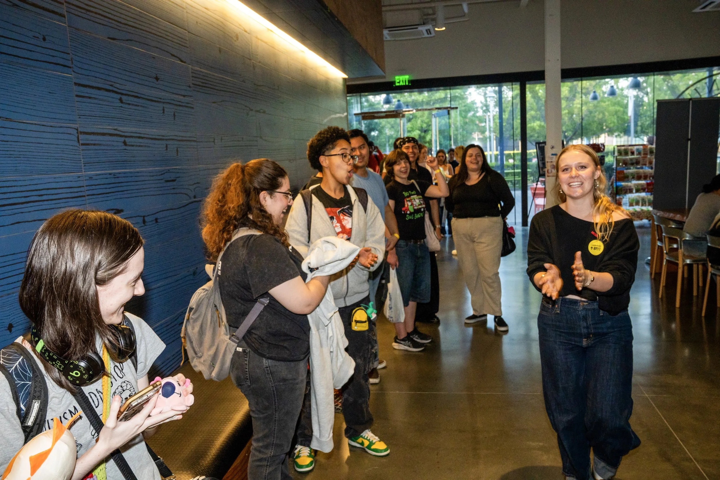 Group of smiling people inside a building, socializing, some holding phones and backpacks, front woman in black top smiling and gesturing with hands.
