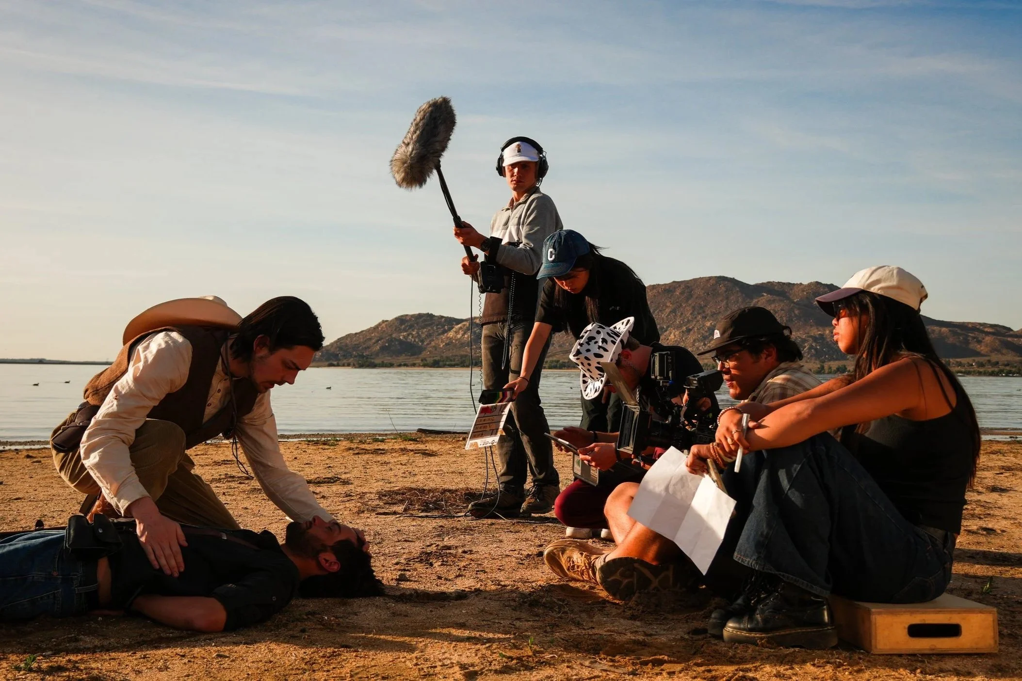 Filmmaking crew filming a scene outdoors near a body of water with mountains in the background. A man with long hair and a beard lies on the ground, while others are crouched or standing around him, some using clipboards and cameras. One person holds a large windscreen-covered microphone.